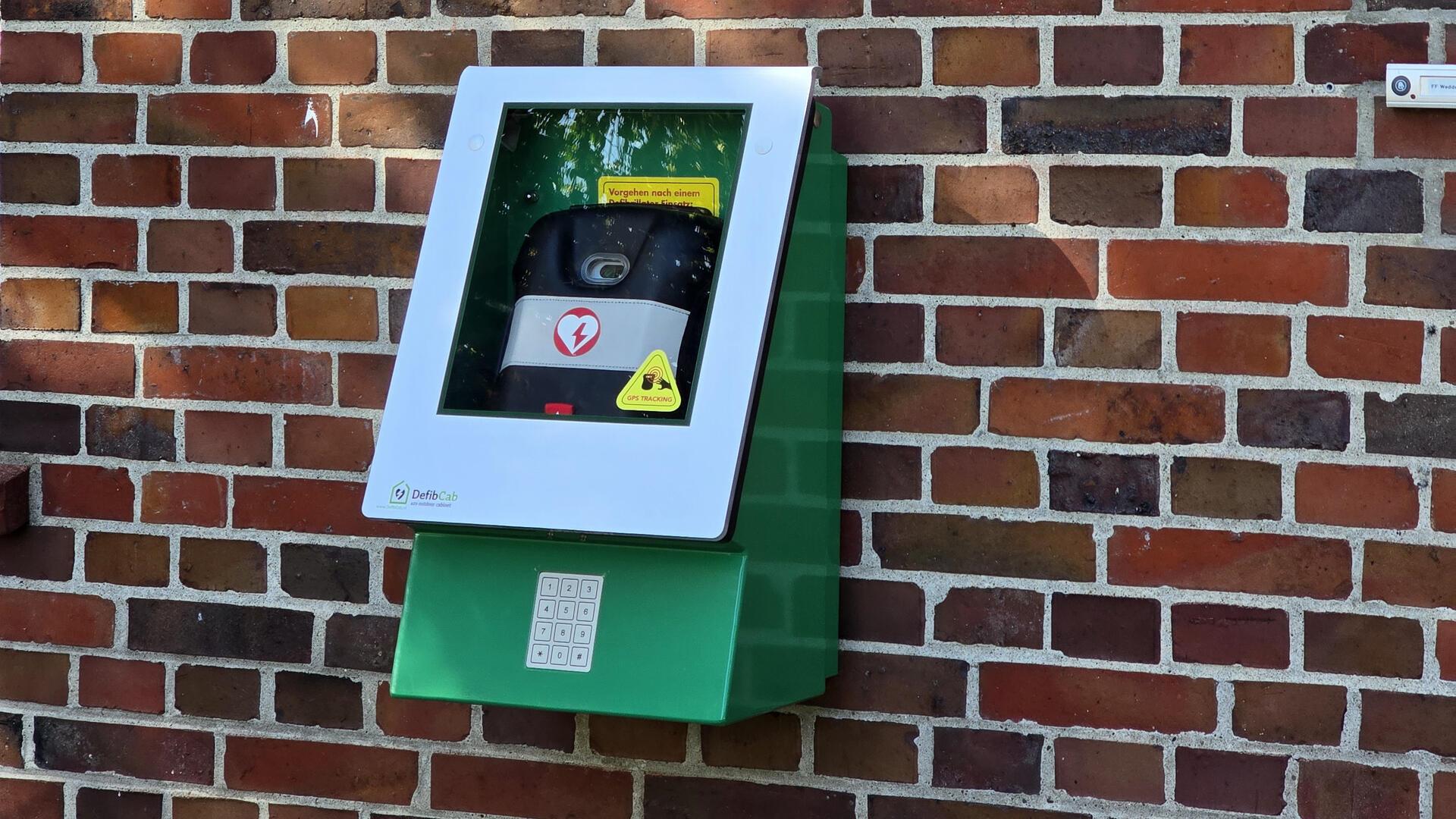 Hinter einer Scheibe in einem grünen Kasten ist ein Defibrillator sichtbar.