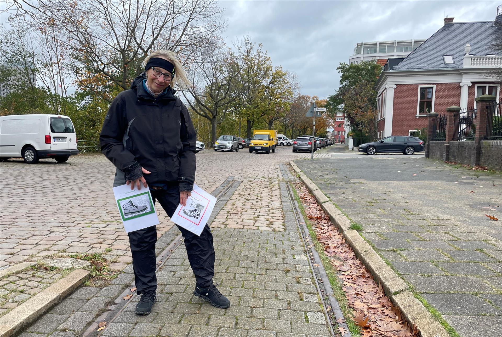 Eine Frau, die auf einer Straße steht. Sie hält zwei Zettel in den Händen. Darauf sind jeweils Stiefel zu sehen, einmal rot und einmal grün umrandet.