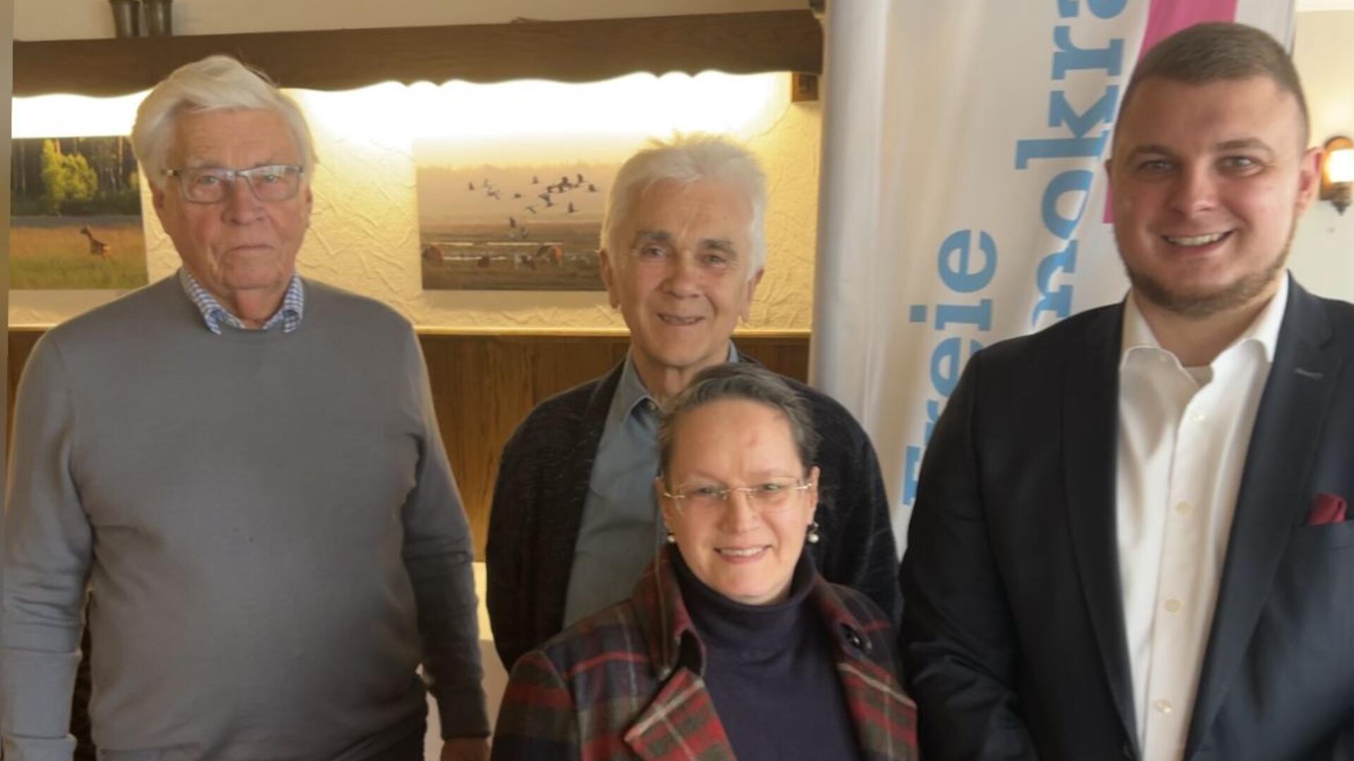 Vier Personen stehen in einem Innenraum vor einer Wand mit beleuchteten Bildern und einer Fahne mit der Aufschrift „Freie Demokraten“. Die Gruppe besteht aus zwei älteren Männern, einer Frau mit Brille und einem jüngeren Mann im Anzug.