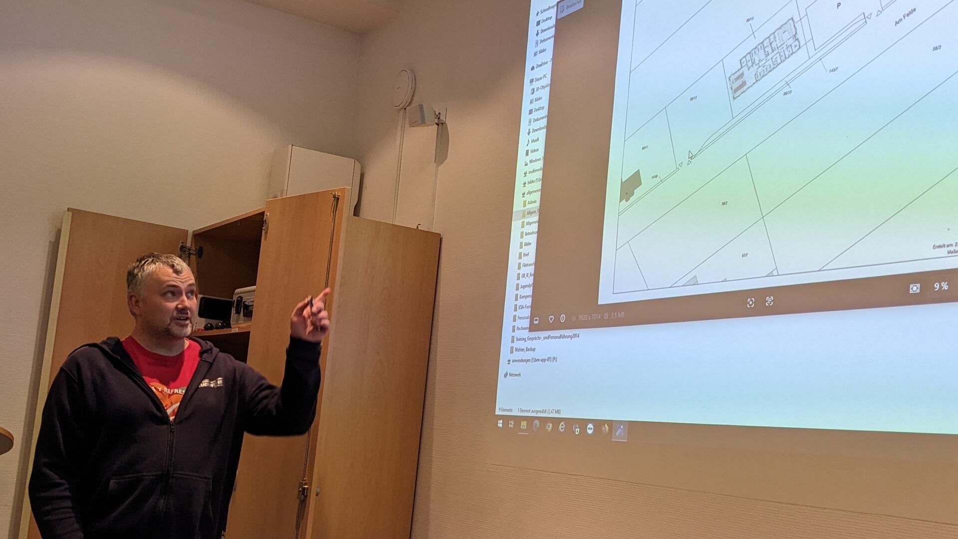 Ein Mann steht vor einer Wand und zeigt auf eine projizierte Bauplan-Zeichnung. Er erklärt etwas und gestikuliert. Im Hintergrund ist ein offener Schrank sichtbar.