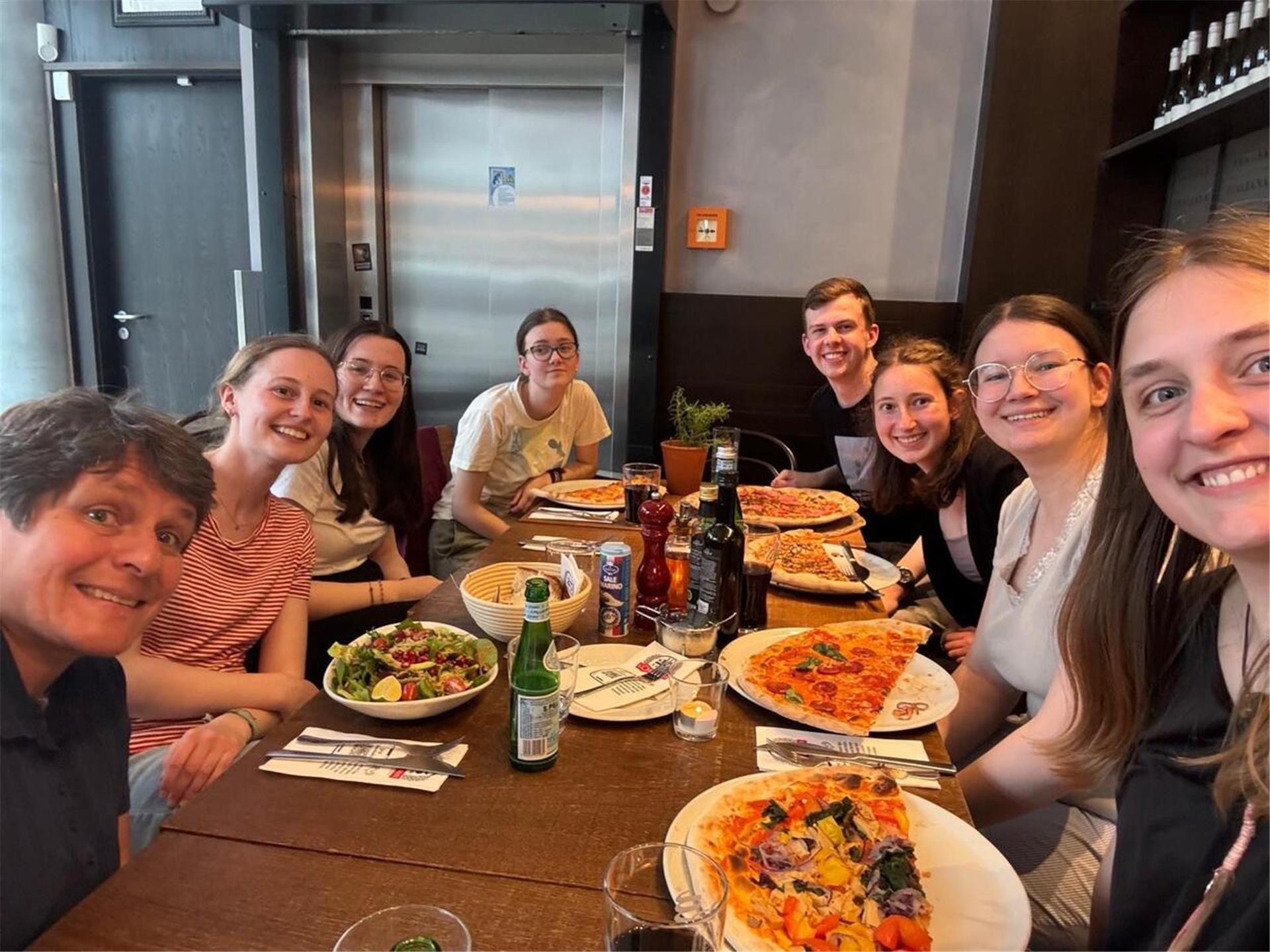 Frauen und Männer sitzen mit Pizza und Salat auf Tellern an einem Tisch und lachen in die Kamera.