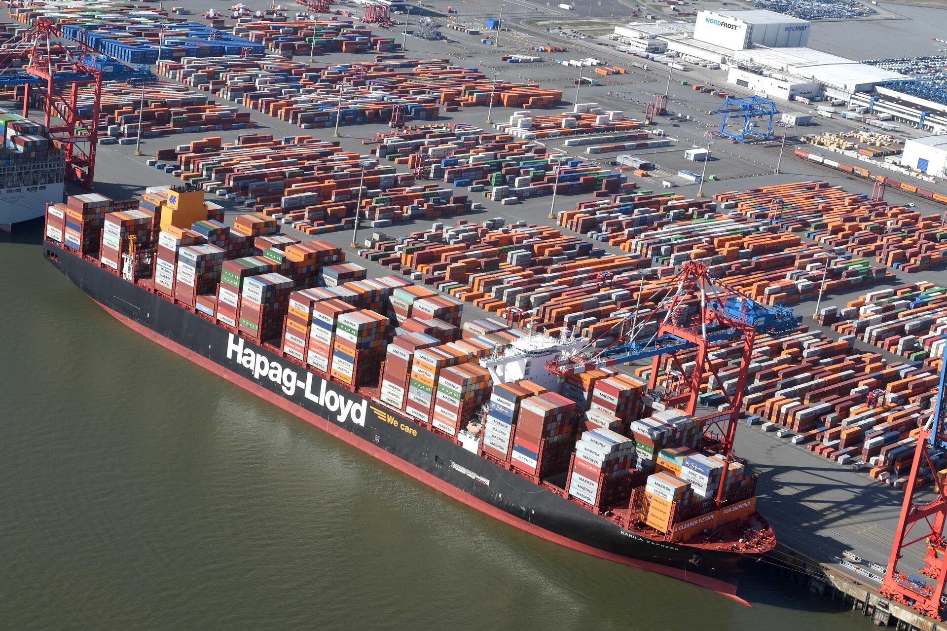 Blick auf einen Hafen, ein großes Containerschiff liegt vor Anker. 