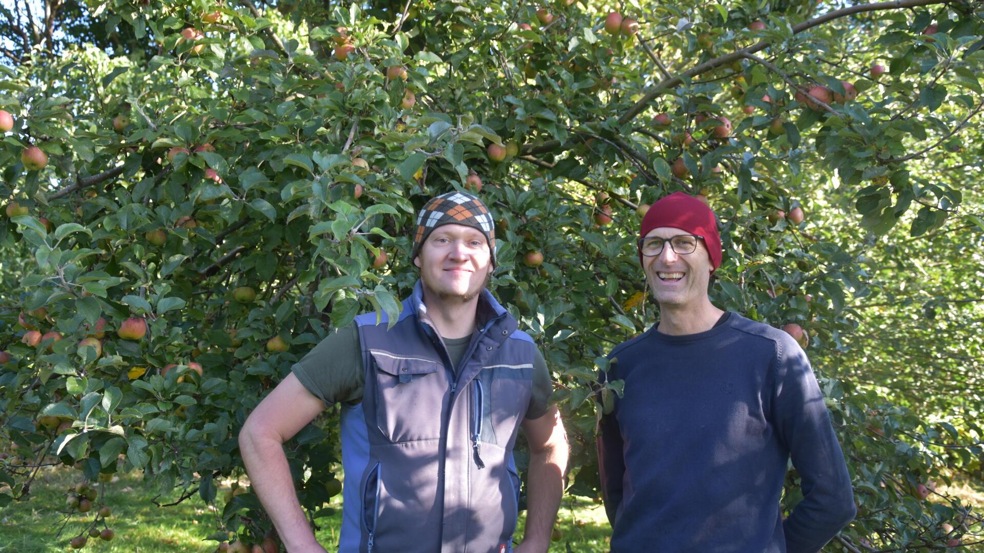 Zwei Männer stehen unter einem Apfelbaum und lachen in die Kamera.
