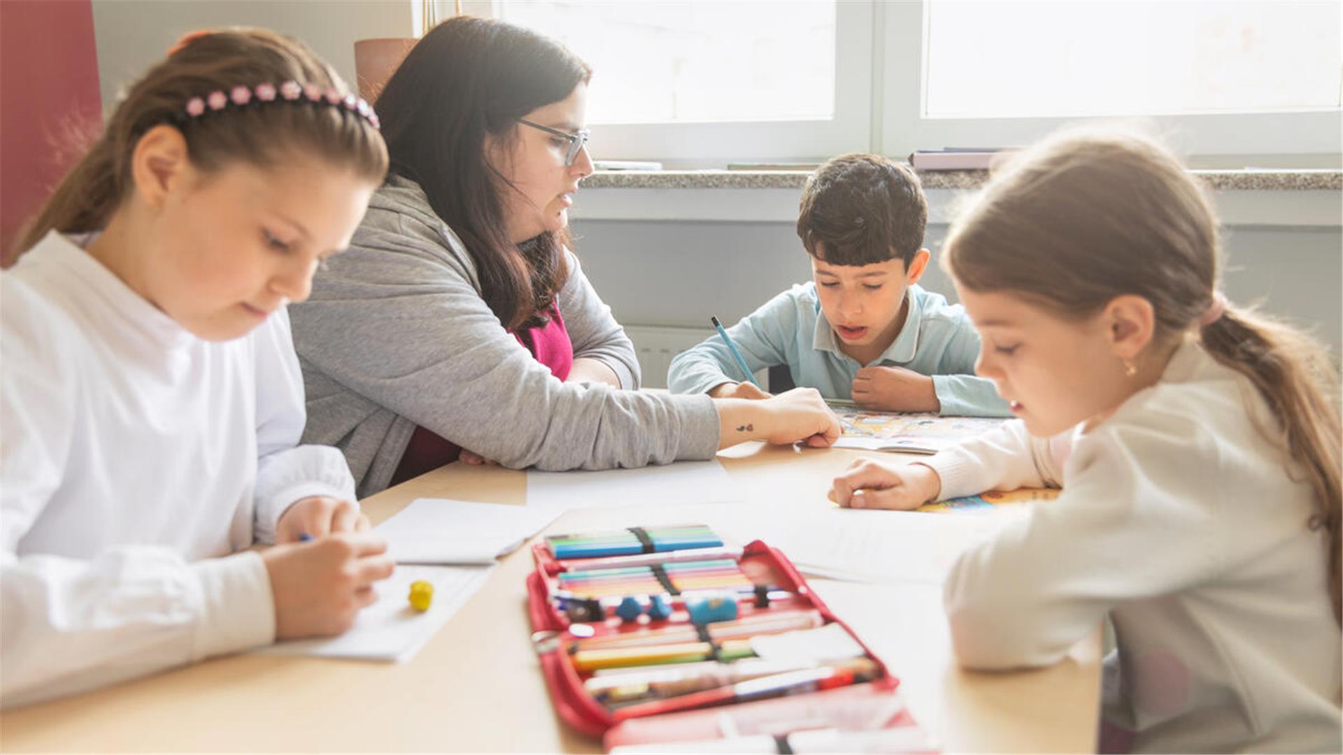 Drei Kinder und eine Frau an einem Tisch. Auf dem Tisch eine Federtasche und verschiedene Blätter.