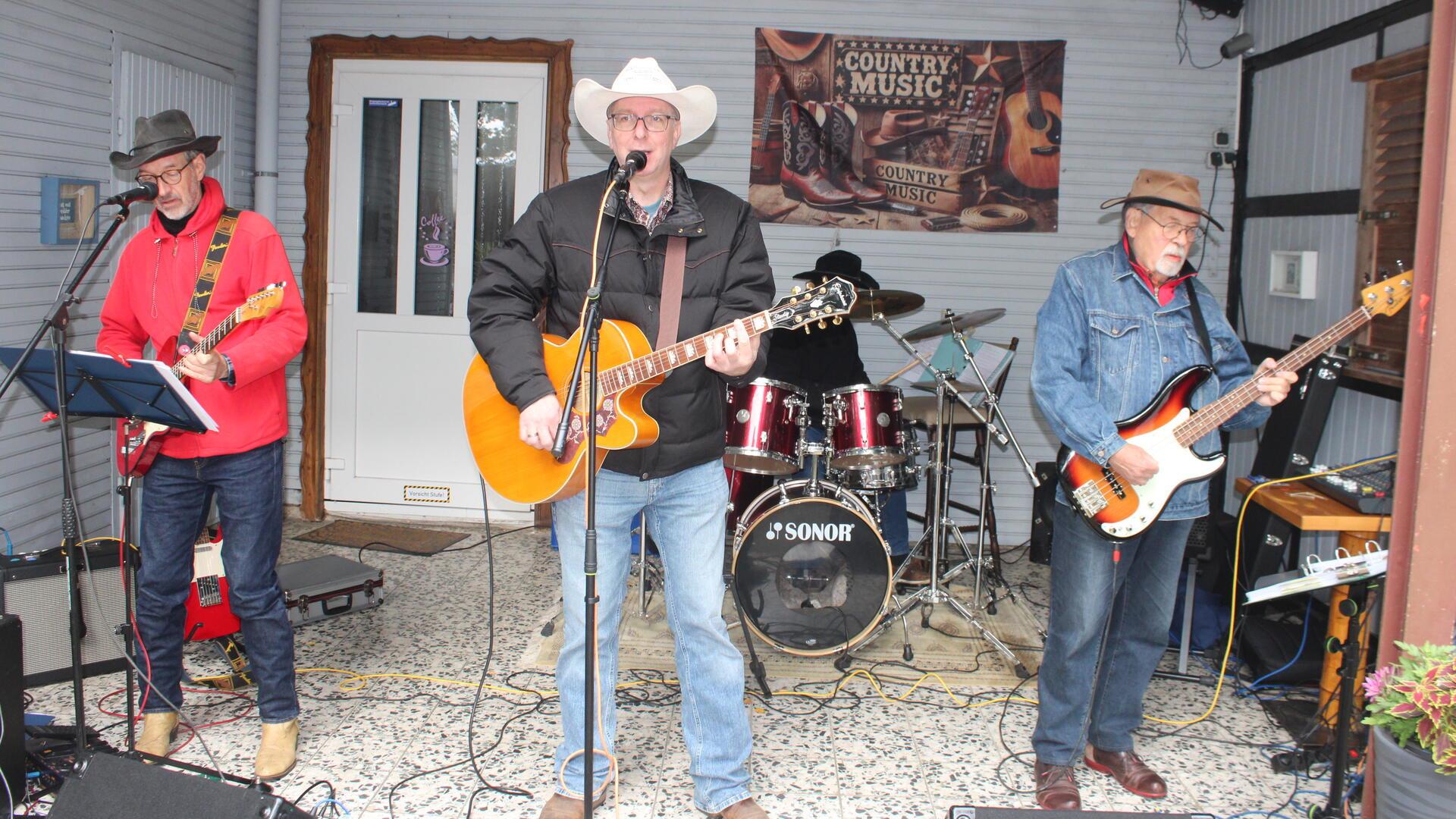 Eine Countryband spielt live auf einer kleinen überdachten Bühne. Drei Musiker mit Gitarren und Cowboyhüten stehen vorne, im Hintergrund sitzt ein Schlagzeuger.