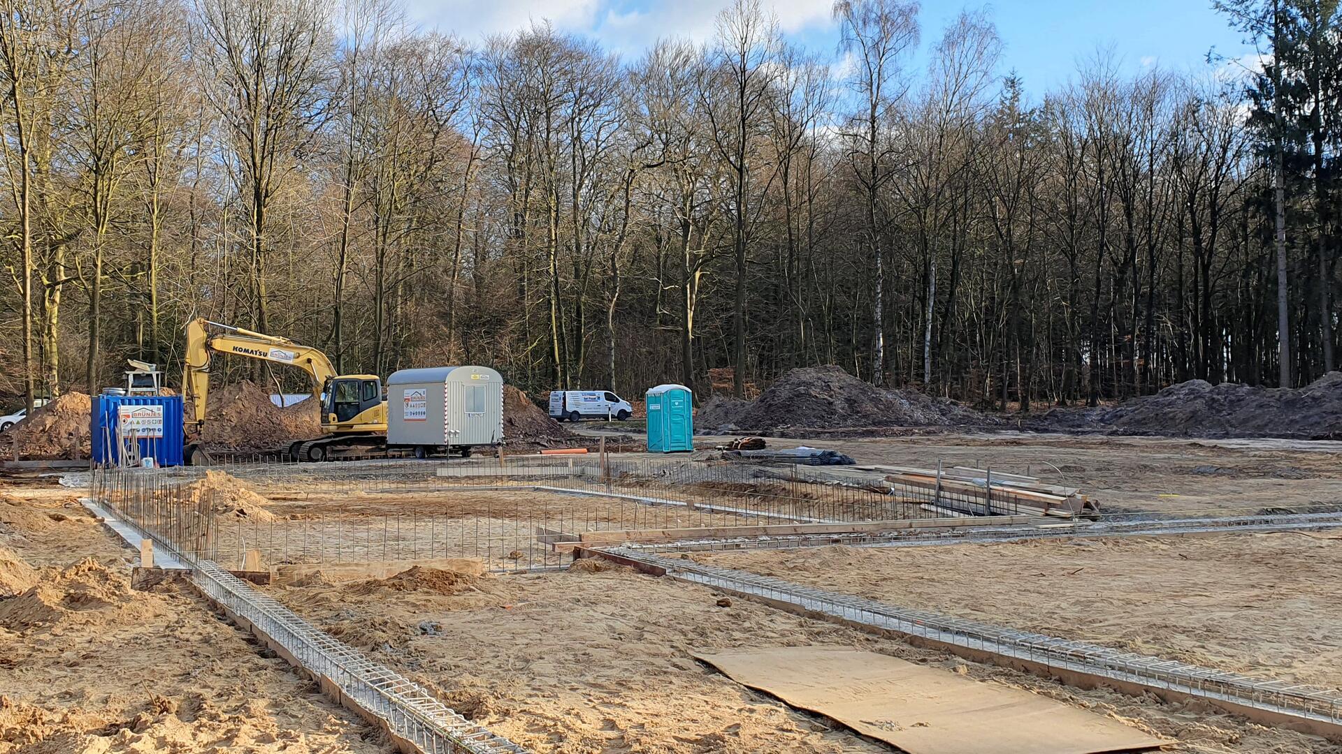 Baustelle im Wald: Ein Bagger steht zwischen Erdhügeln, umgeben von Baucontainern und mobilen Toiletten. Die Fundamente eines neuen Gebäudes sind bereits vorbereitet. Im Hintergrund erstreckt sich ein dichter Laubwald.