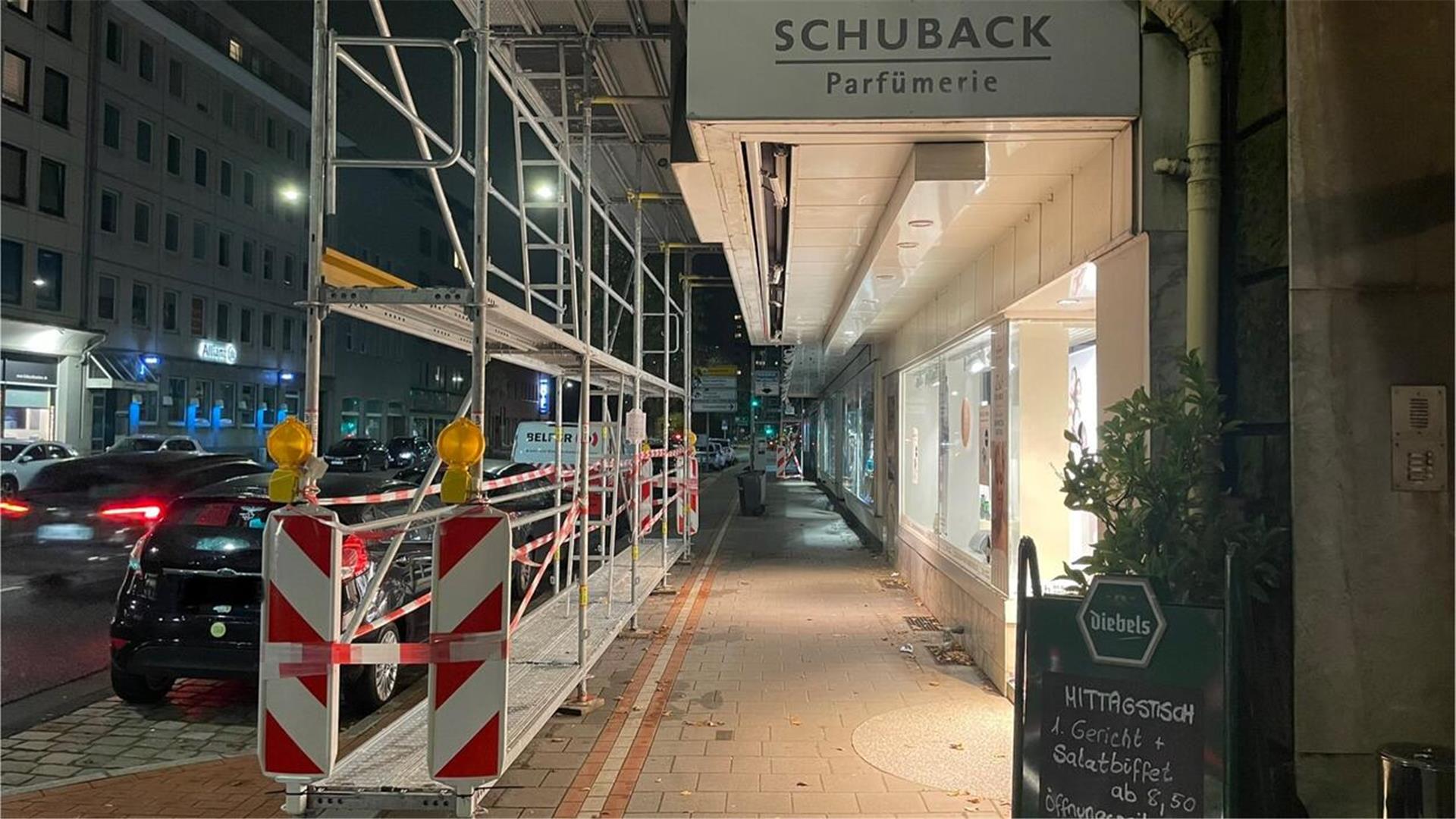 Blick auf einen Fußweg und einen Radweg, auf dem ein Baugerüst steht. Oben im Bild ist der Schriftzug "Schuback Parfümerie" zu sehen. Links fahren Autos eine Straße entlang.