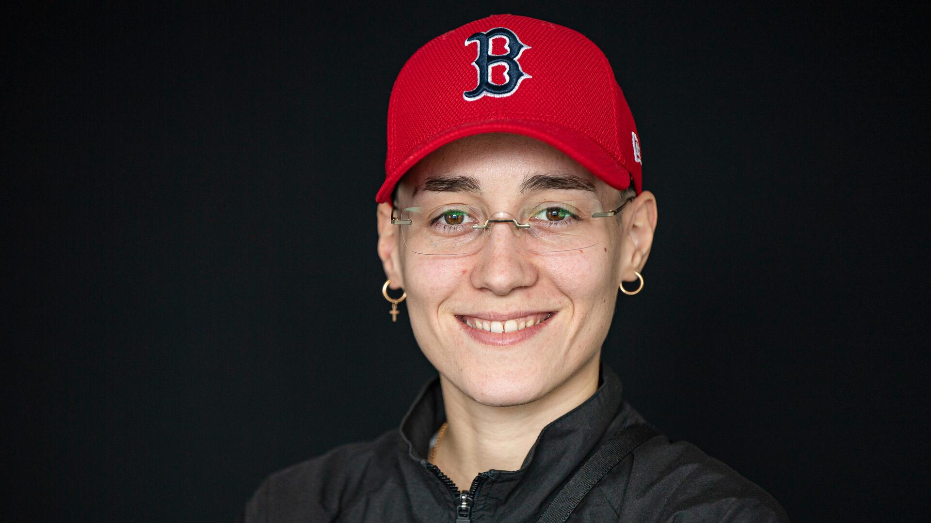 Das Gesicht einer jungen Frau, die Brille, goldene Ohrringe und eine rote Baseballkappe trägt.