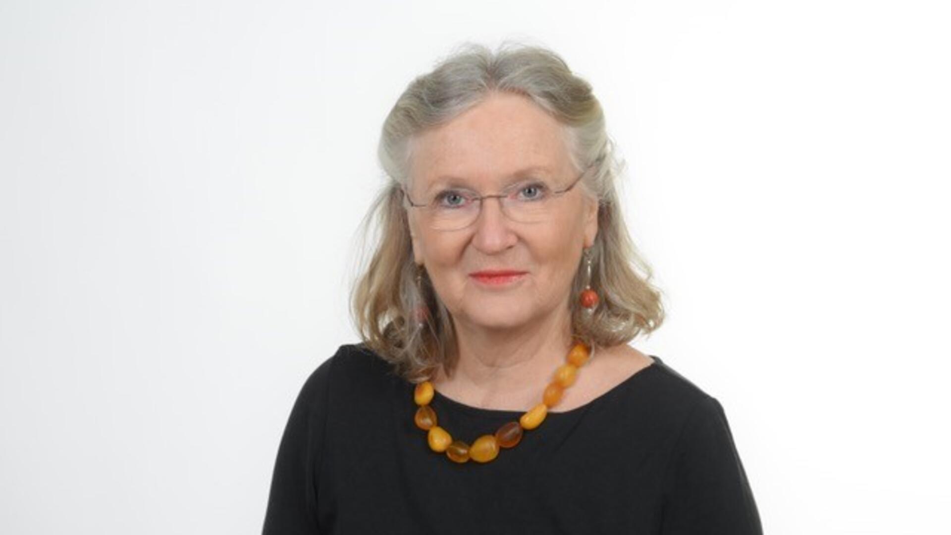 Autorin Monika Kiel-Hinrichsen: graue schulterlange Haare, Brille, ernter Blick. Schwarzes Oberteil, Bernsteinkette