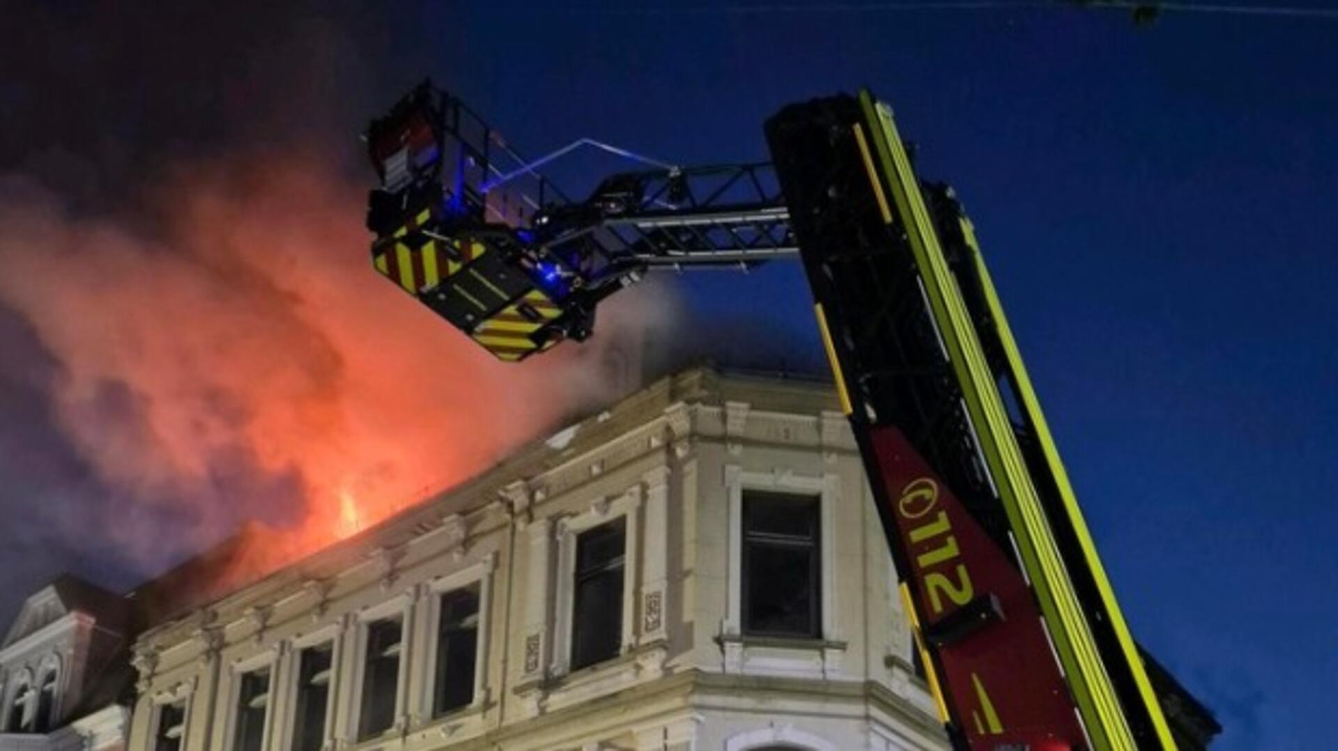 Flammen schlagen aus einem Haus im Hintergrund. Im Vordergrund ist die Drehleiter der feuerwehr zu sehen.