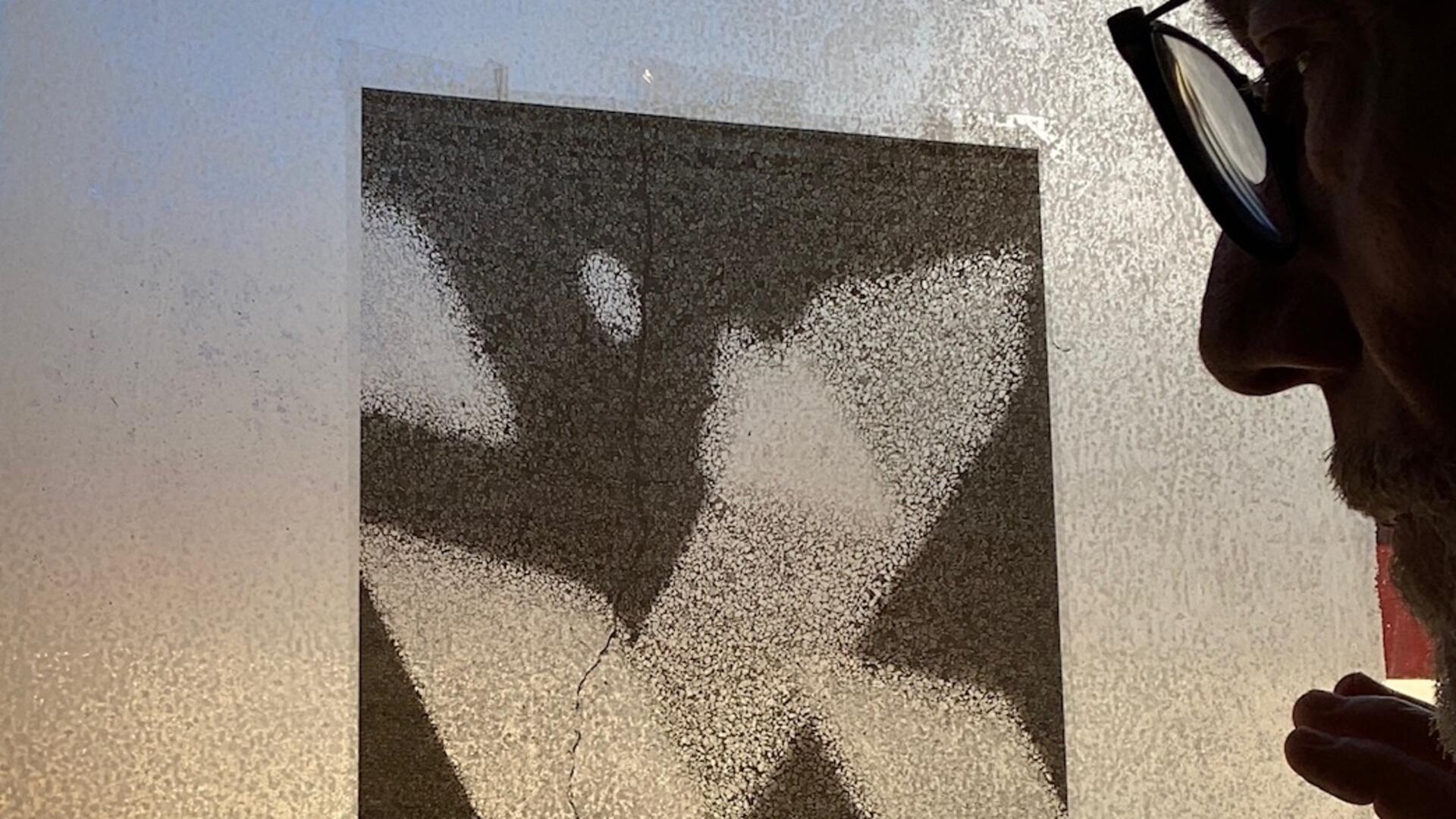 Ein Mann mit Brille betrachtet eine abstrakte Schwarz-Weiß-Grafik, die an einer mattierten Glasscheibe befestigt ist. Das Licht fällt durch das Glas und betont die Struktur des Bildes sowie das nachdenkliche Profil des Mannes.