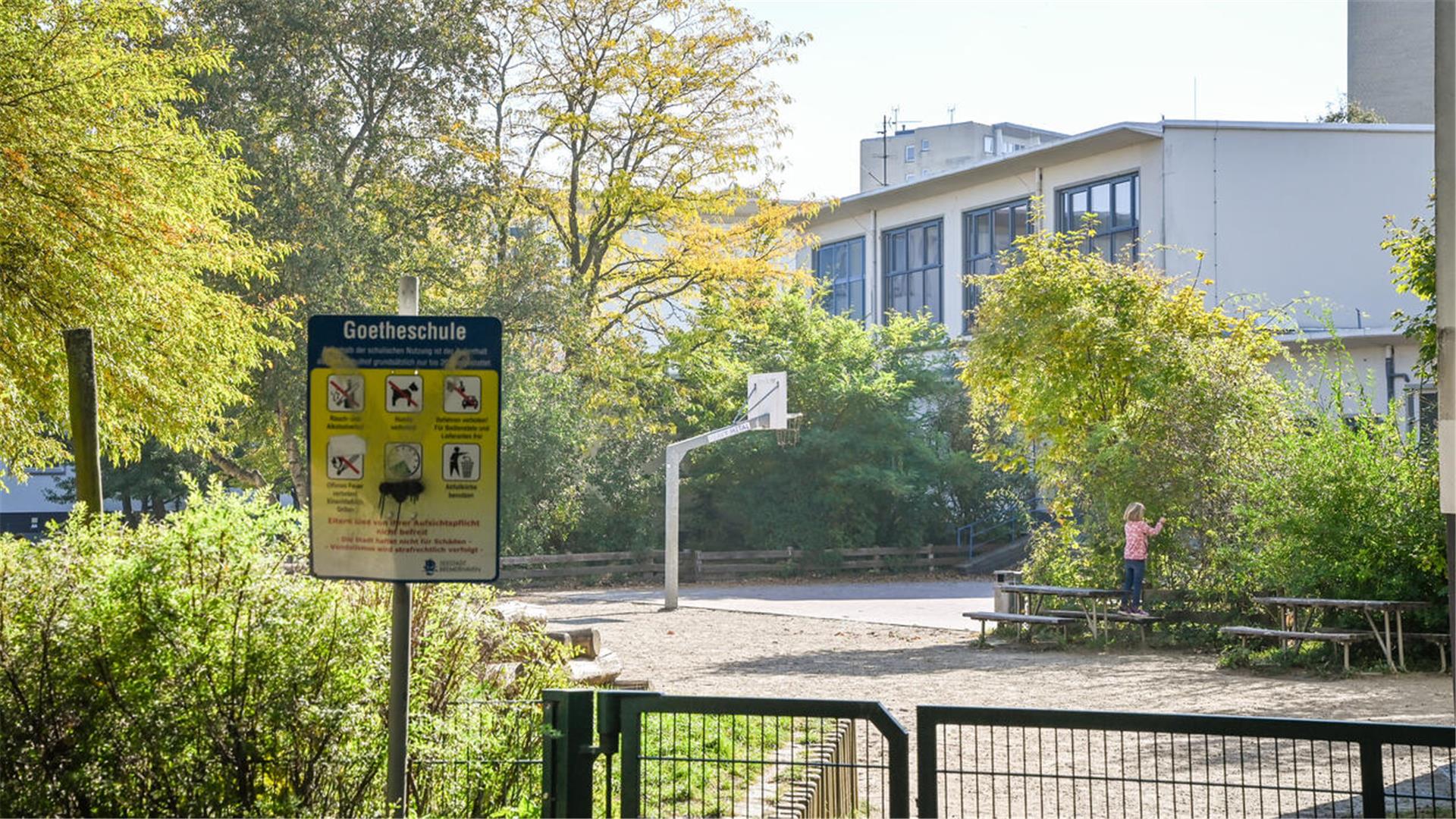 Blick über einen grünen Mattenzaun auf einen Schulhof mit vielen Büschen und einem Basketballkorb. Im Vordergrund steht ein Spielplatz-Schild mit der Aufschrift Goetheschule und sechs Piktogrammen.