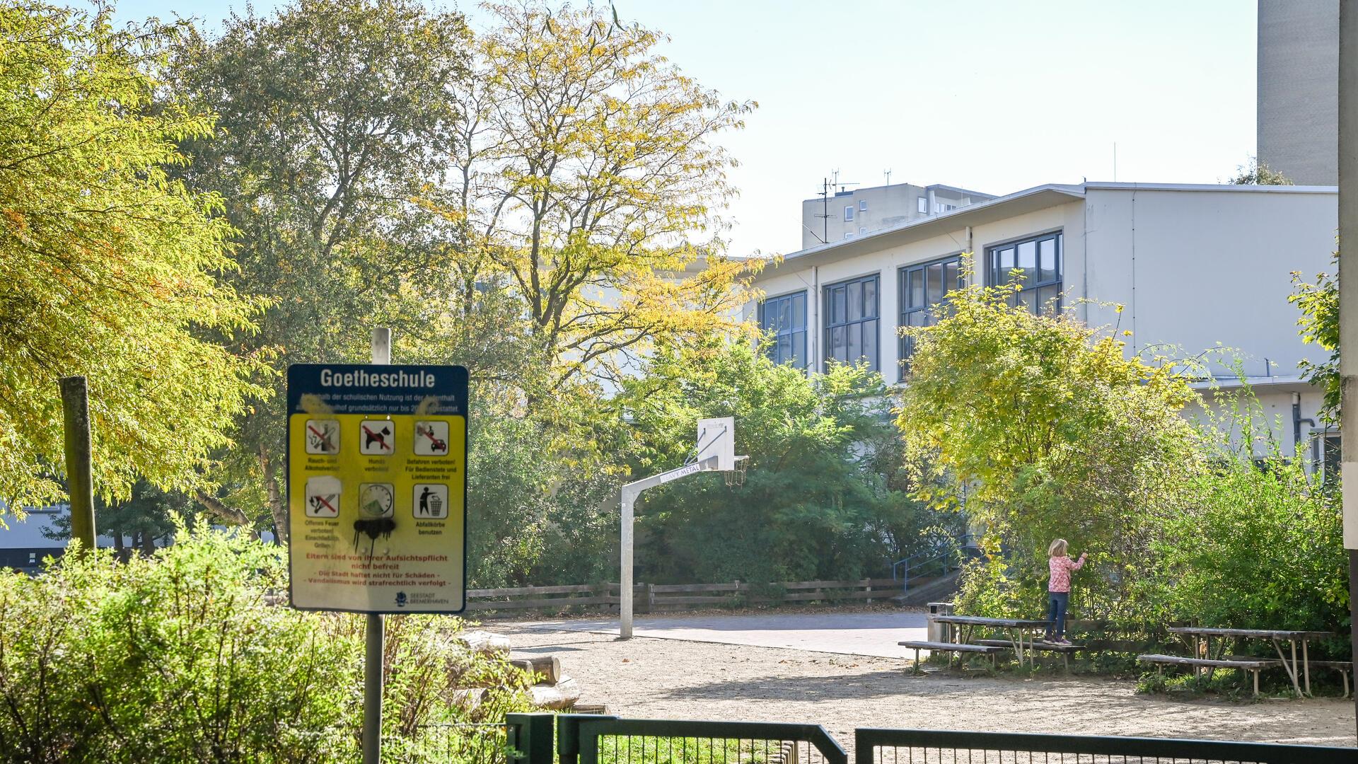 Blick über einen Zaun auf einen Schulhof mit Bänken und einem Basketballkorb