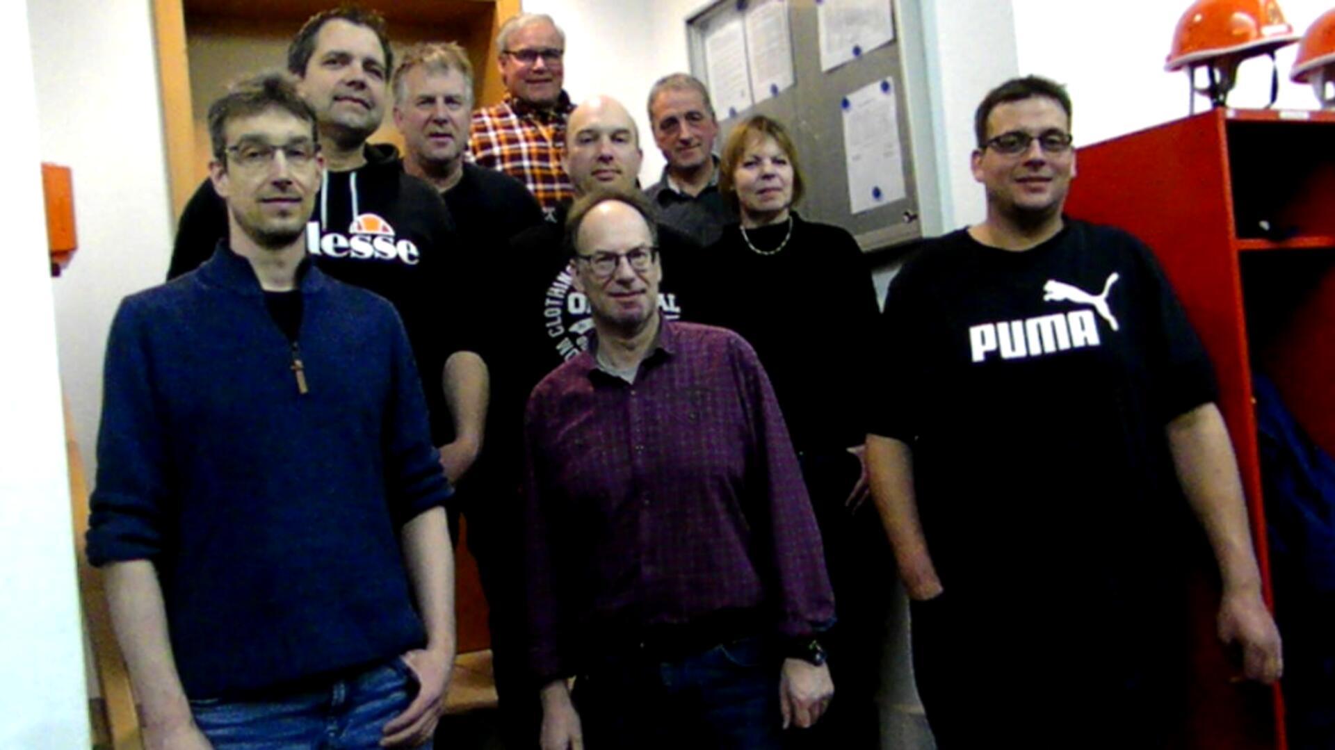 Eine Gruppe von zehn Personen posiert für ein Foto in einem Innenraum. Einige stehen auf einer Treppe, während andere davor stehen. Die Personen tragen überwiegend legere Kleidung, darunter Pullover, T-Shirts und Hemden.