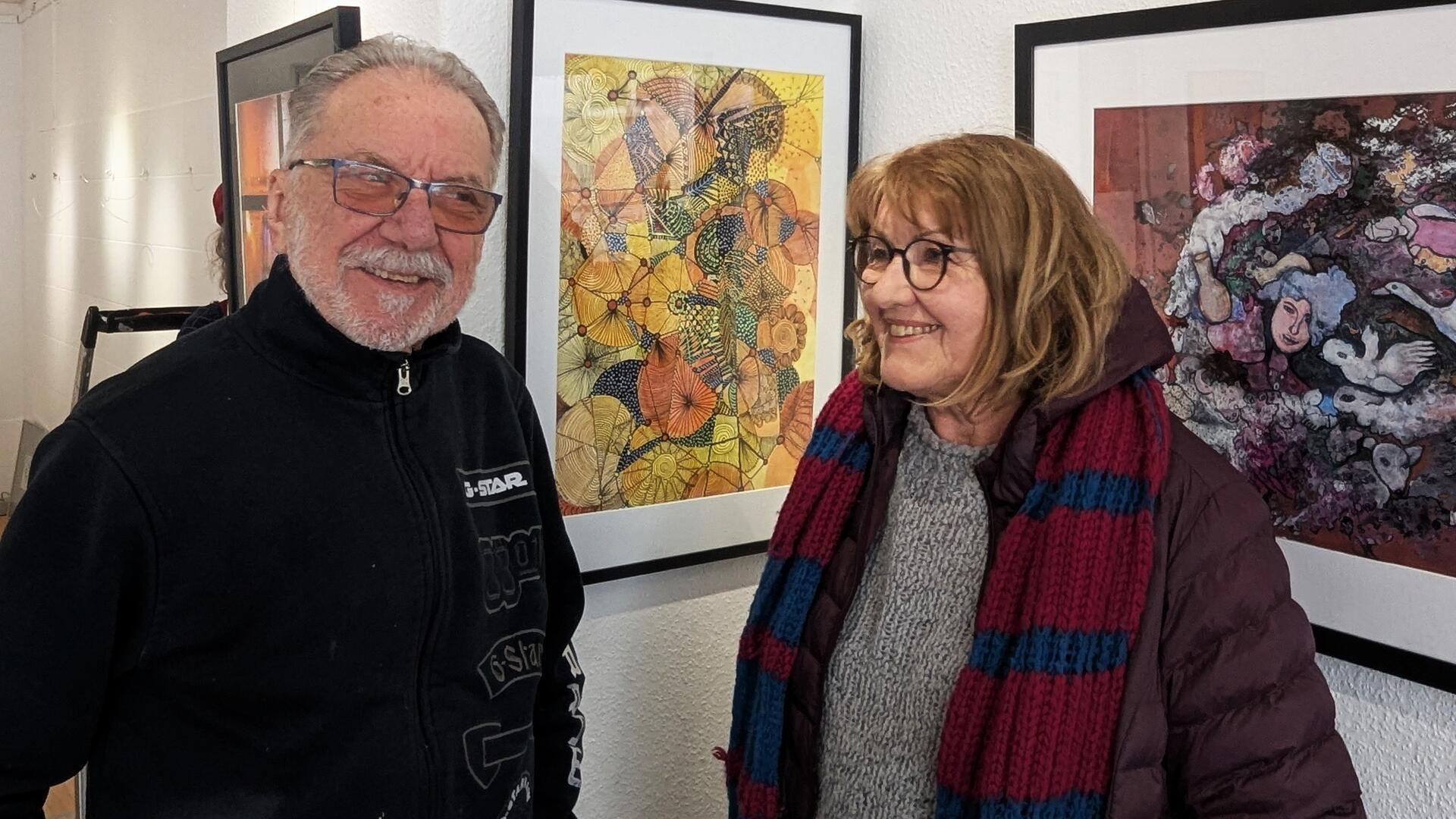 Zwei Personen stehen lächelnd vor gerahmten Kunstwerken in einer Galerie. Links ein älterer Mann mit Brille und schwarzer Jacke, rechts eine Frau mit Brille, brauner Jacke und gestricktem Schal. Im Hintergrund sind farbenfrohe Kunstwerke zu sehen.