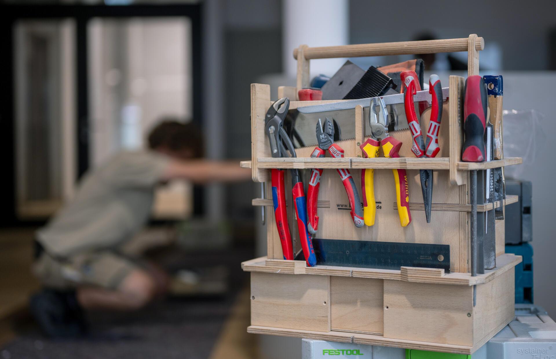 Ein Werkzeugkasten steht auf dem Tisch, während im Hintergrund ein Handwerker ein Möbelstück zusammenbaut.  Die Konjunkturumfrage der Handwerkskammer Bremen zeigt einen Rückgang im Geschäftsklimaindex. Trotz dieser negativen Entwicklung bleibt die Stimmung unter den Betrieben verhalten optimistisch.