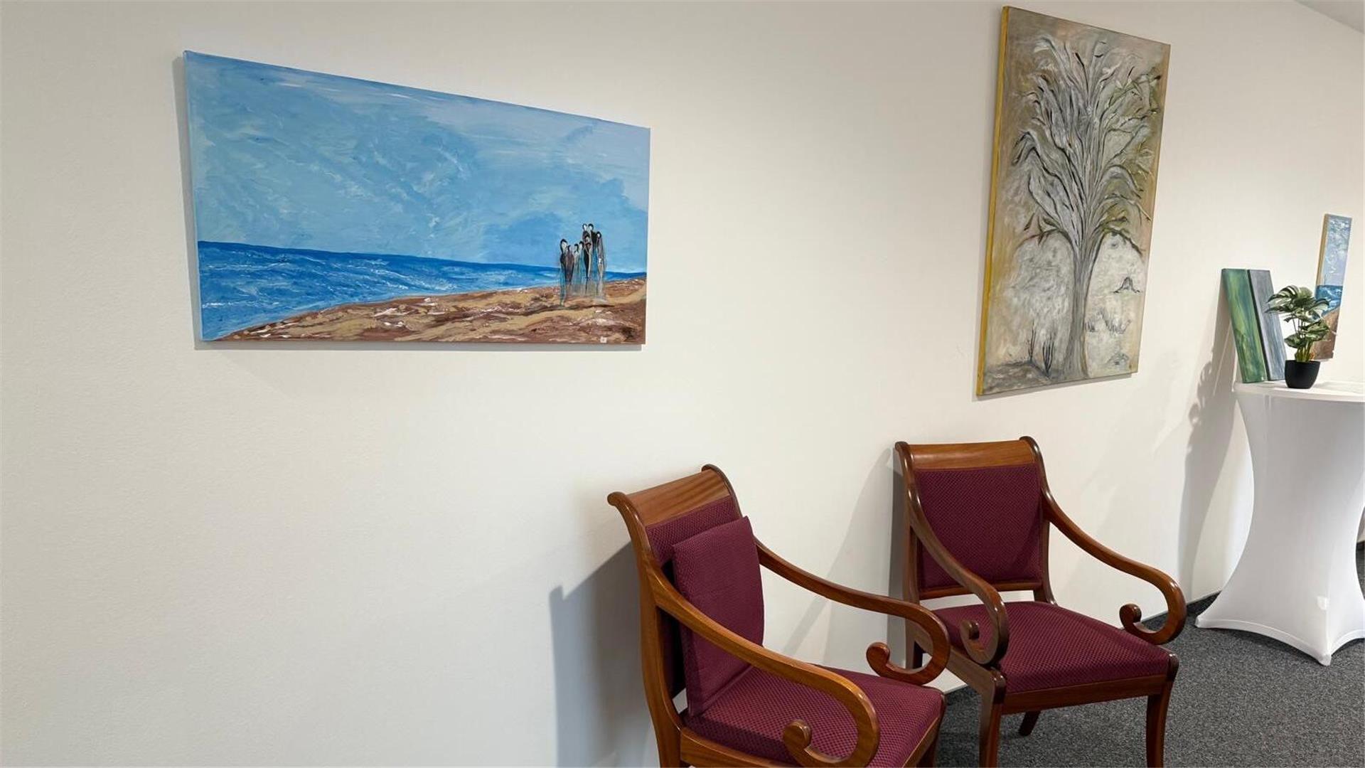 Kunstwerke hängen an einer Wand, davor stehen zwei Stühle. Die Künstlerin Käthe Goltz stellt 27 farbenfrohe Werke im Amtsgericht Zeven aus.