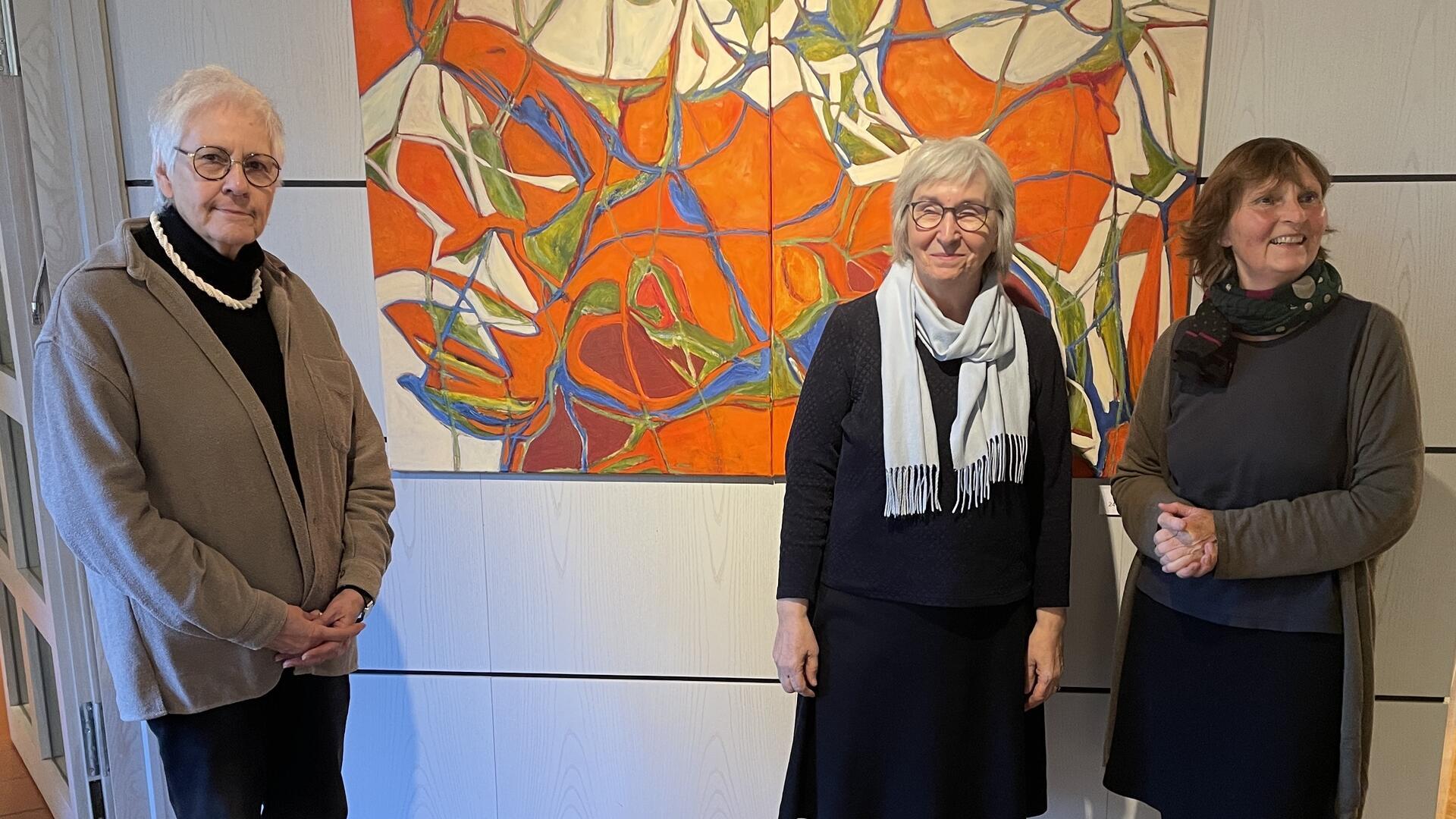 Drei Frauen stehen vor einem farbenfrohen, abstrakten Gemälde mit dominanten Orange-, Blau- und Grüntönen.