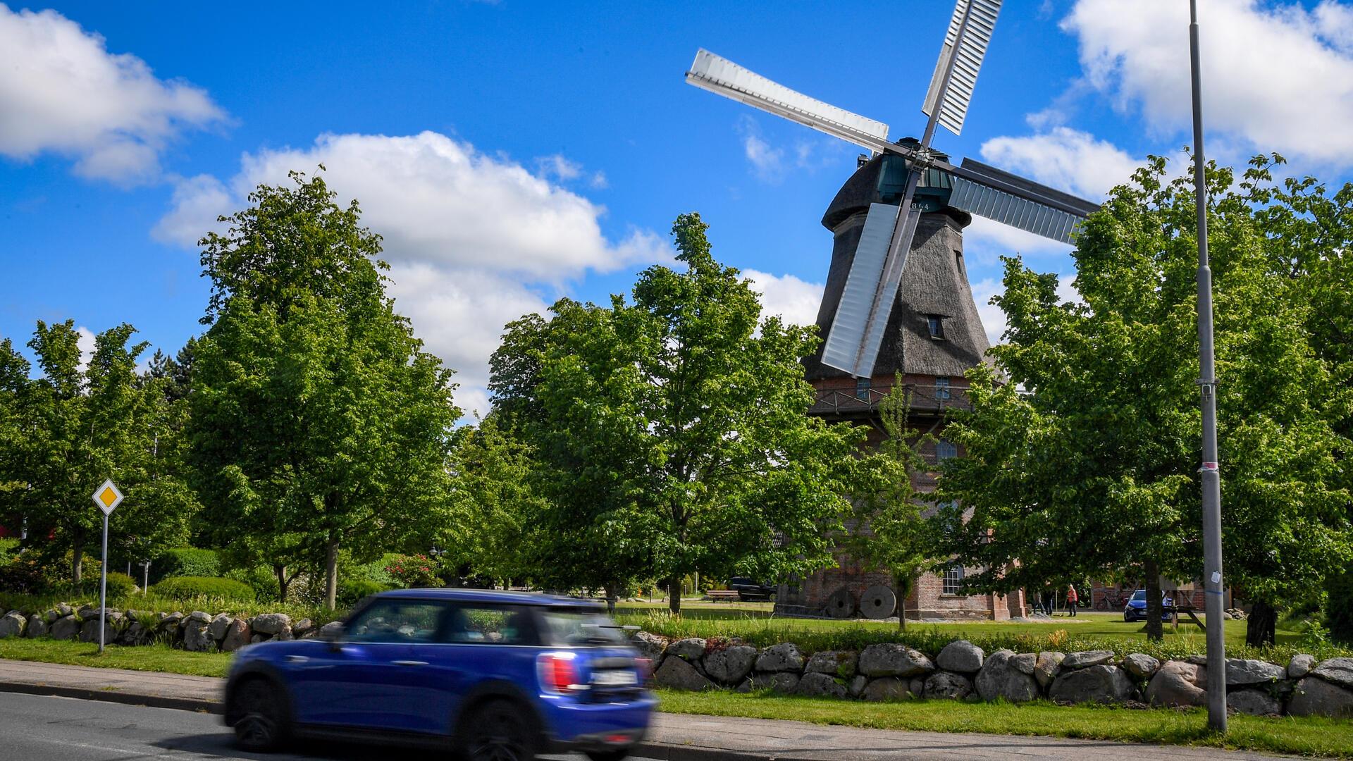 Eine Mühle steht in einem Dorf mitten im Grünen. Im Vordergrund eine Straße, auf der ein blaues Auto vorbeifährt.