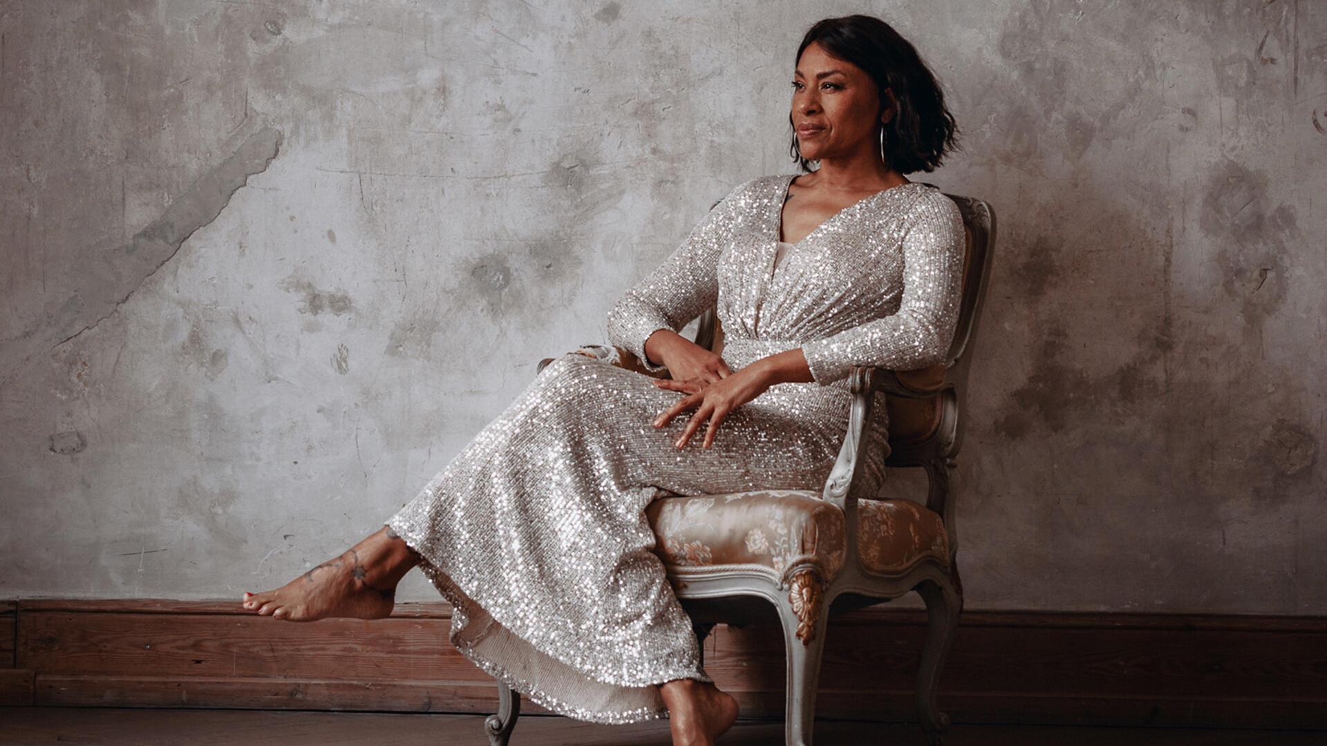 Eine dunkelhäutige Frau in einem schimmernden Kleid sitzt auf einem Stuhl.
