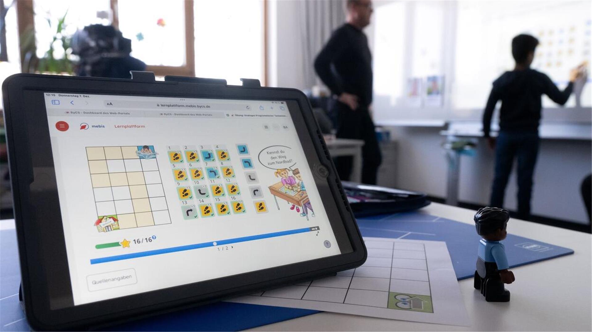Ein Tablet in einem Klassenzimmer, im Hintergrund ein Schüler und ein Lehrer an der Tafel.