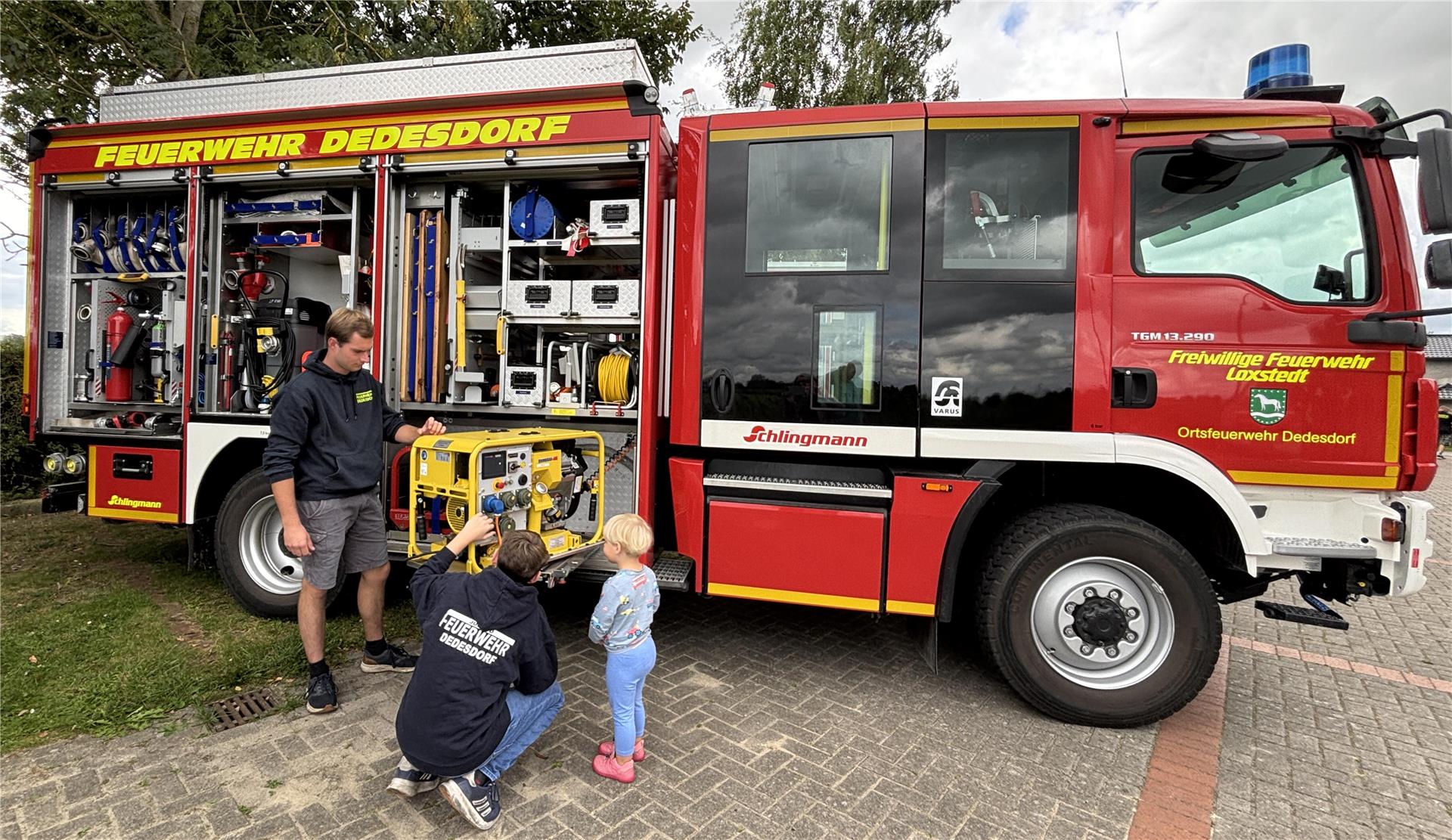 Zwei Feuerwehrleute der Ortsfeuerwehr Dedesdorf zeigen einem kleinen Kind technische Geräte am geöffneten Einsatzfahrzeug. Der Wagen ist mit Ausrüstung beladen und gut sichtbar beschriftet.