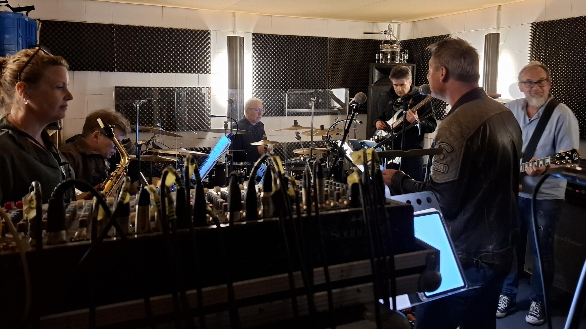 Mehrköpfige Band bei der Probe in einem schallgedämmten Raum mit Schlagzeug, Gitarren, Mischpult und Mikrofonen; konzentrierte Atmosphäre im Tonstudio.