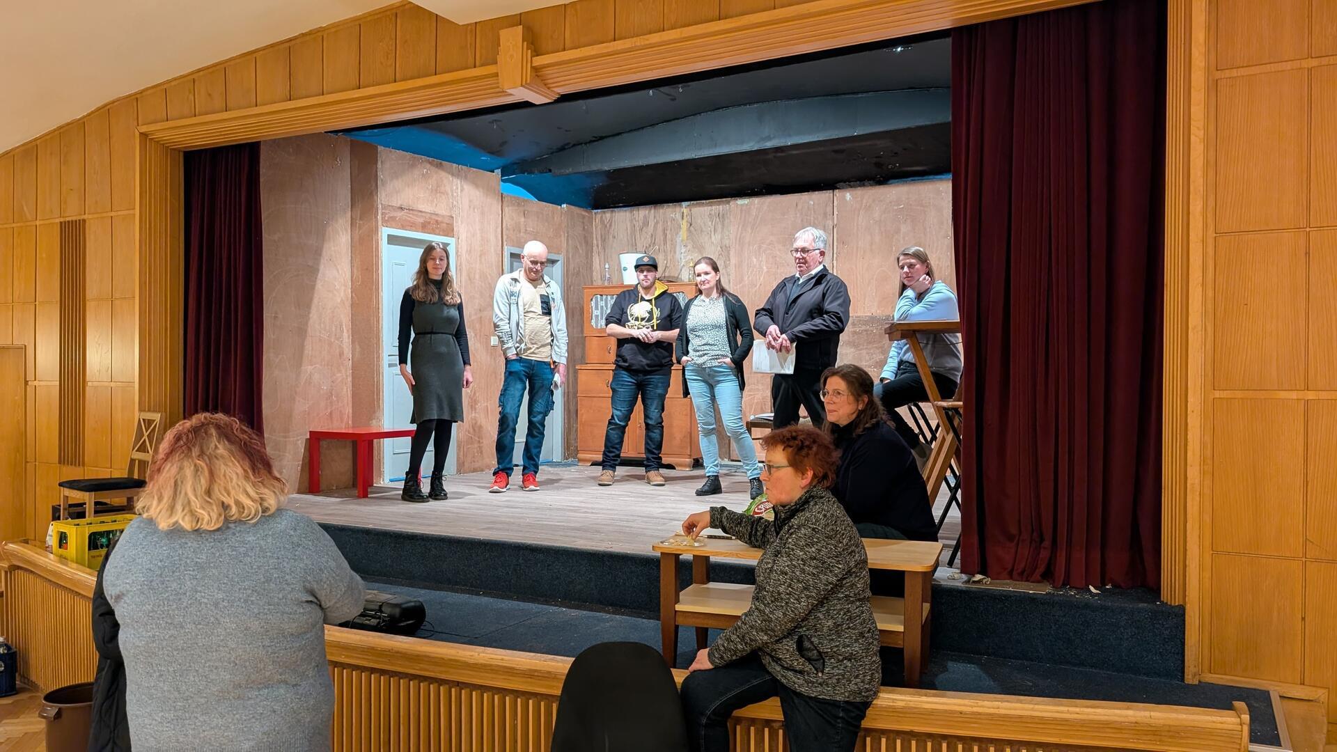 Sechs Personen stehen auf einer Theaterbühne, während drei davor sitzen. Die Aufnahme zeigt die Theaterprobe.