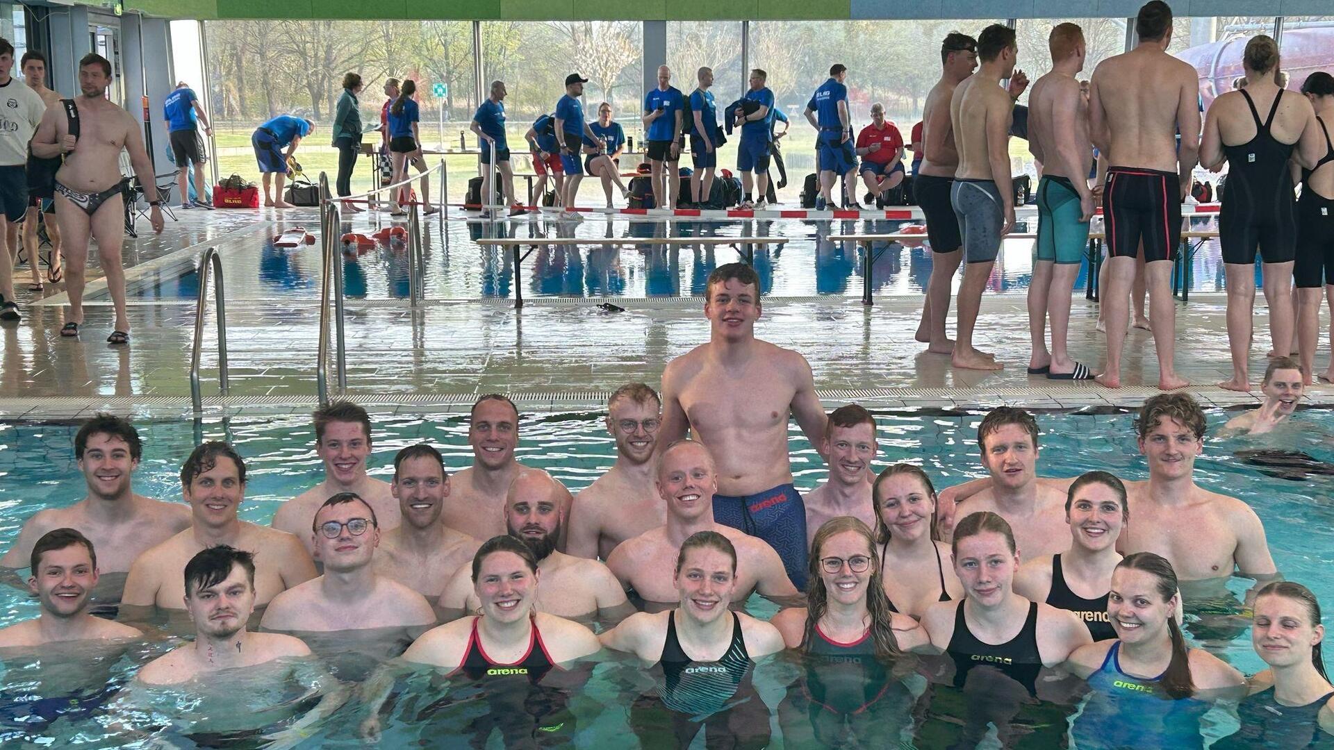 Gruppenfoto von rund 25 jungen Erwachsenen der DLRG Hagen im Bremischen, die in Badekleidung im Schwimmbecken stehen und lächeln; im Hintergrund weitere Wettkampfteilnehmende am Beckenrand einer Schwimmhalle.
