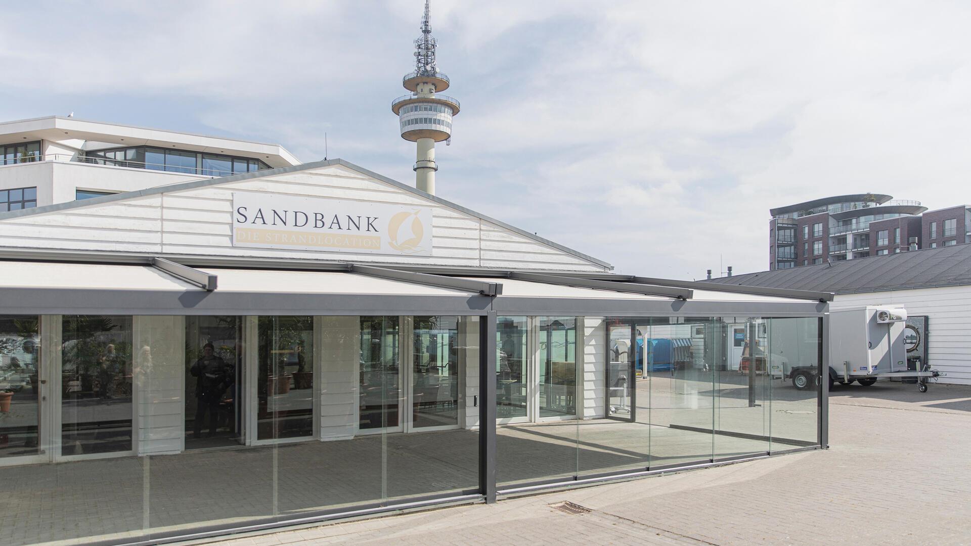 Blick auf ein Gebäude. Die Sandbank präsentiert sich mit neuer Terrasse und Glasfront.
