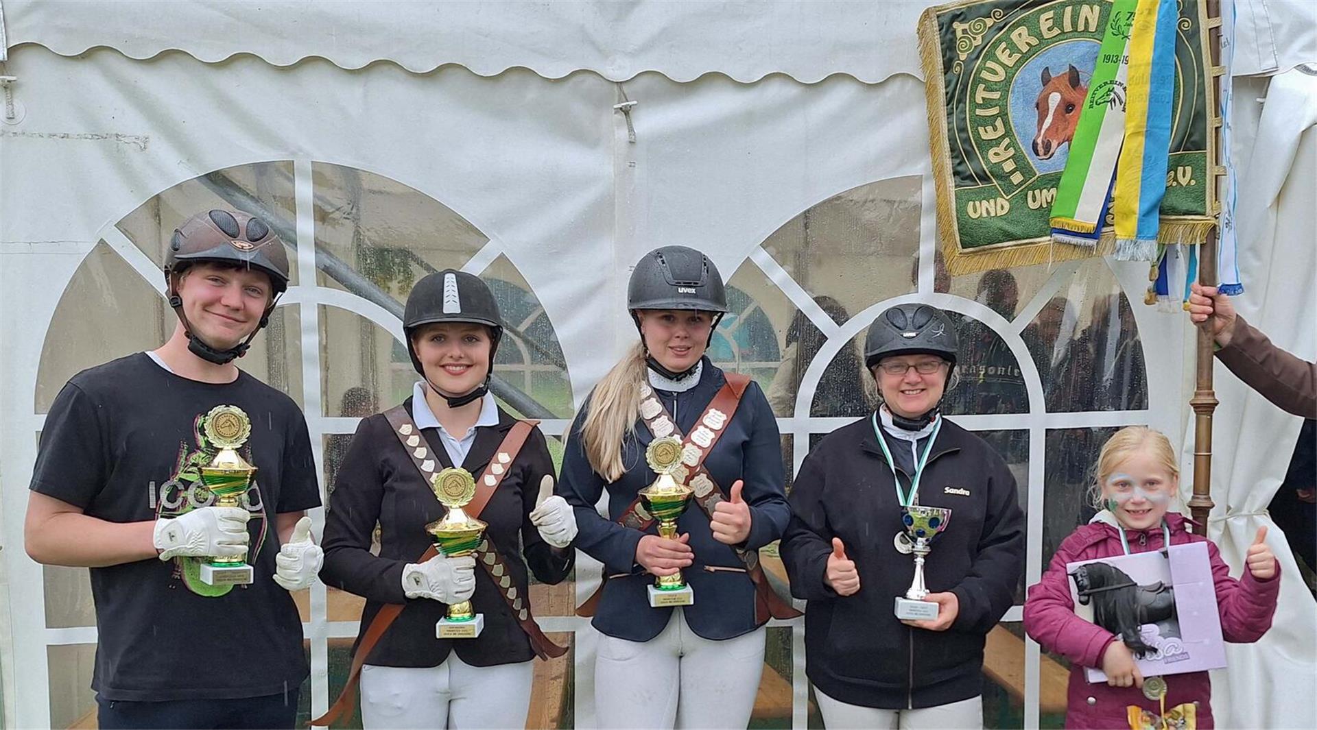 Gruppenfoto der erfolgreichen Teilnehmerinnen und Teilnehmer beim Ringreiten des Reitvereins Hagen, mit Pokalen, Schärpen, Reithelmen und Vereinsfahne vor einem weißen Festzelt.