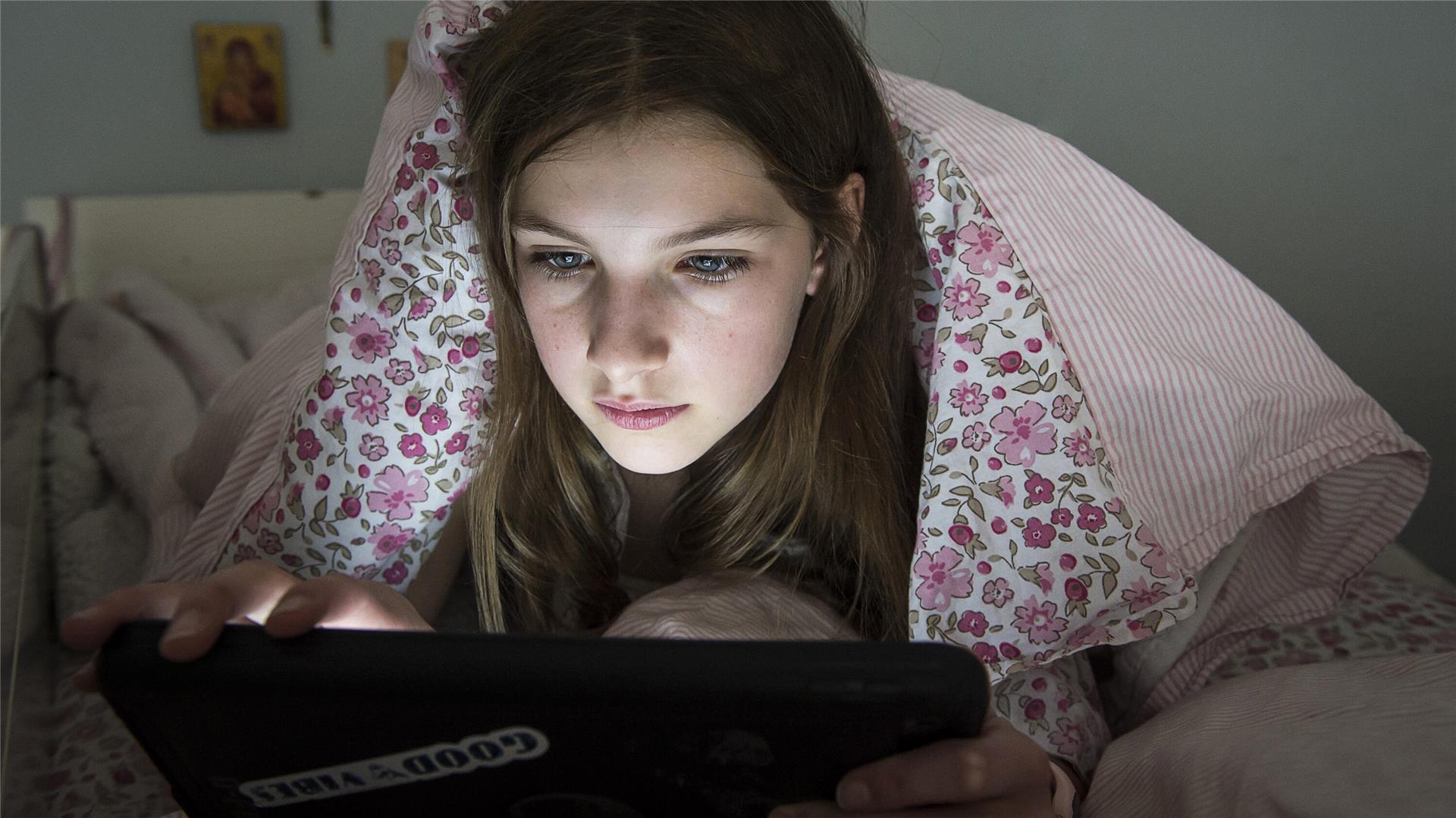 Ein Mädchen liegt auf dem Bett unter der Decke und schaut auf ein Tablet.