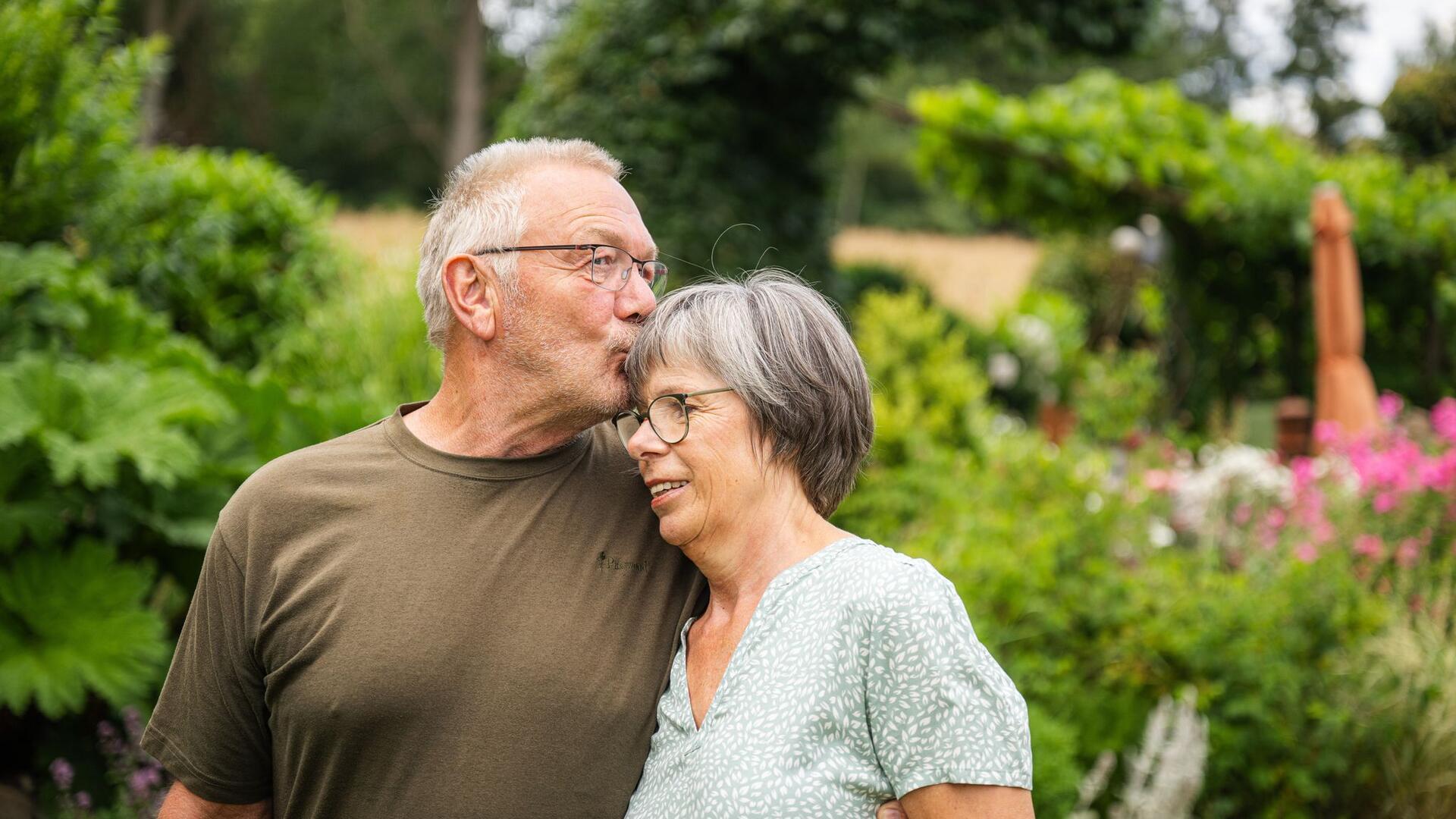 Ein älteres Paar steht in einem Garten. Der Mann küsst die Frau auf die Stirn.