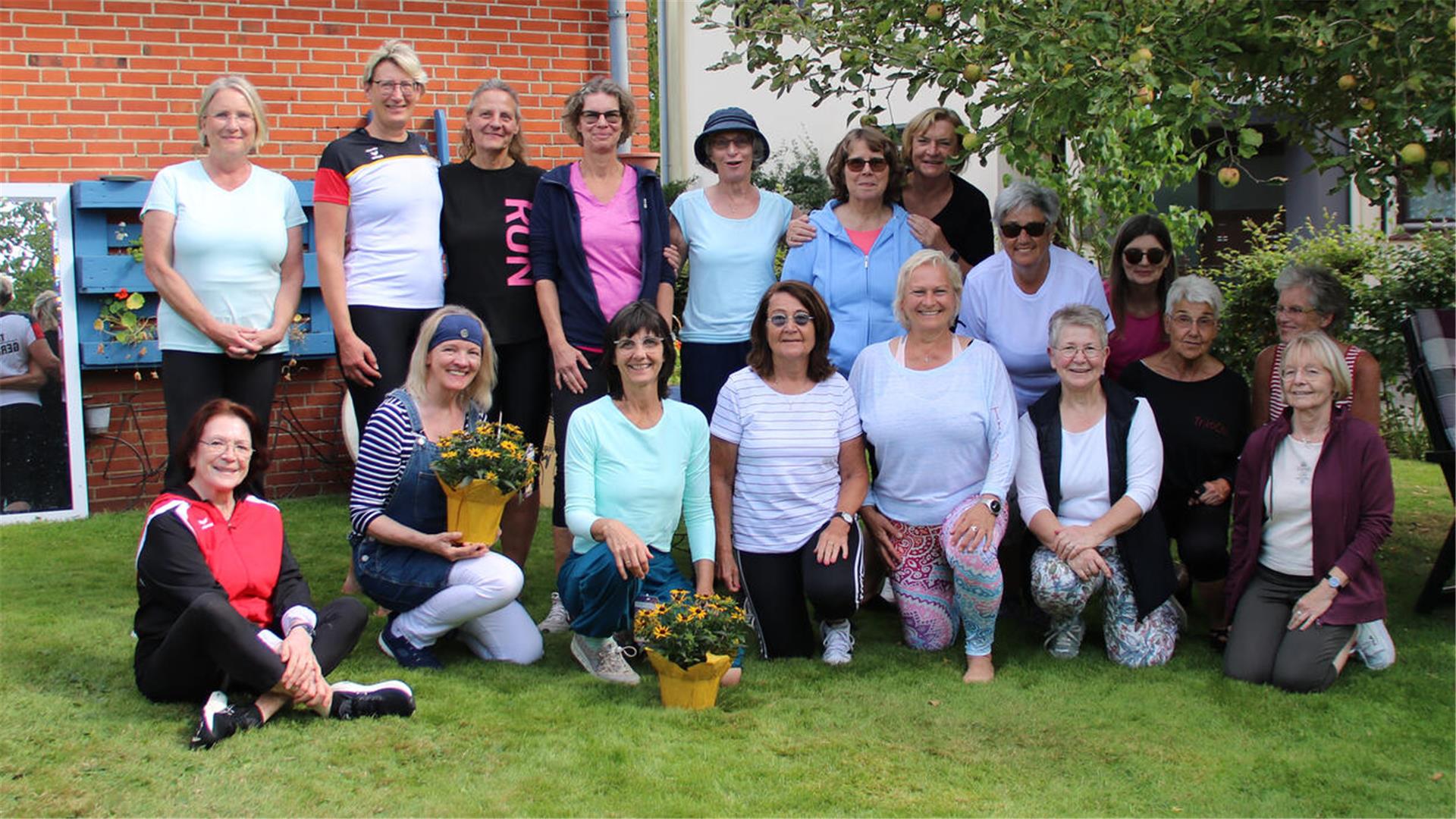 Eine Gruppe von Frauen unterschiedlichen Alters posiert fröhlich im Garten für ein Gruppenfoto. Einige knien oder sitzen im Gras, zwei halten Blumenschalen in den Händen. Im Hintergrund sind ein Apfelbaum, eine Hauswand und ein Spiegel zu sehen.