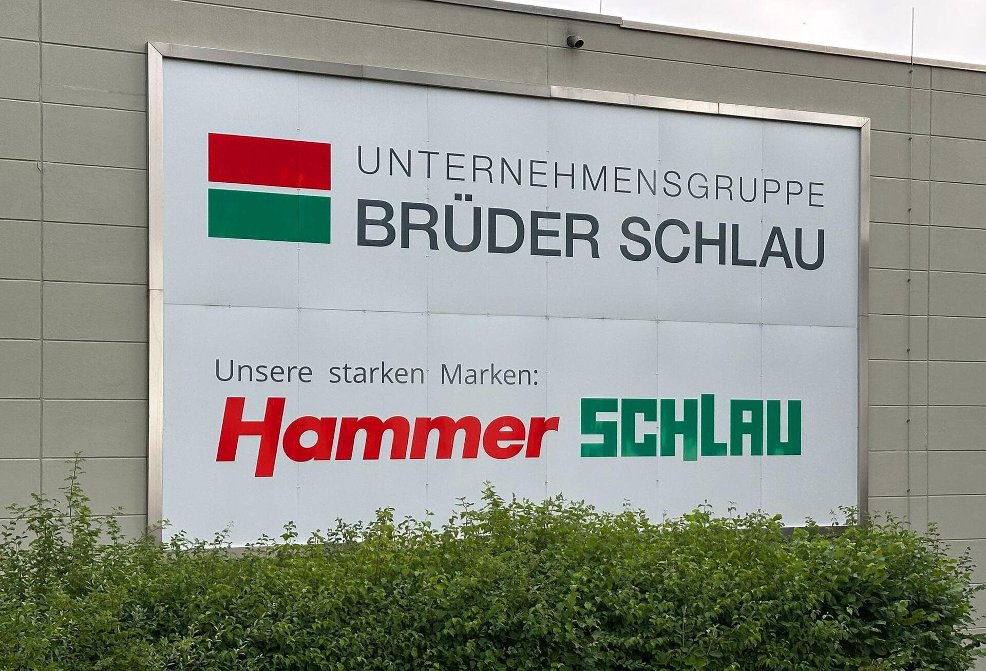 Das Logo der Unternehmensgruppe Brüder Schlau GmbH & Co KG und das des Raumaustatters Hammer ist auf einem Schild zu sehen.