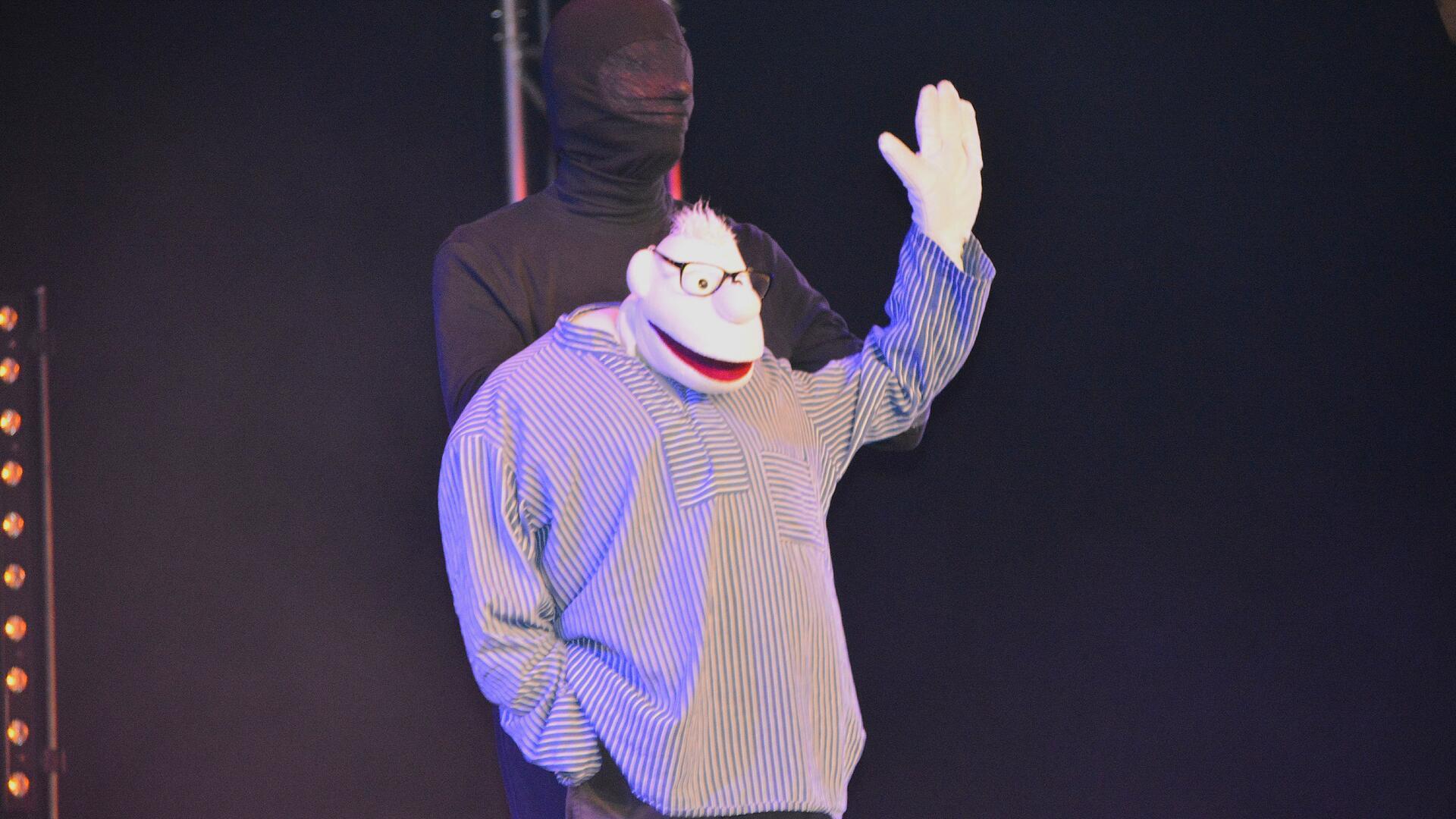 Ein Puppenspieler steht in schwarz gekleidet auf der Bühne und hält eine Stoffpuppe. 