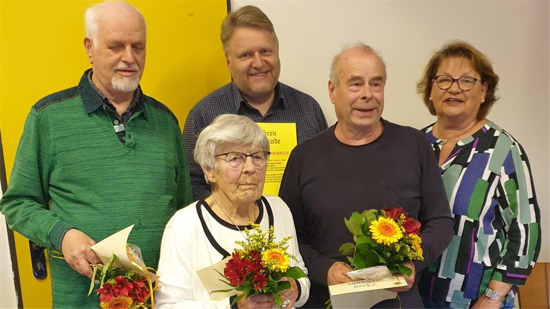 Fünf Personen, drei Männer und zwei Frauen, stehen zusammen. Vier von ihnen halten Blumensträuße und Urkunden in den Händen.