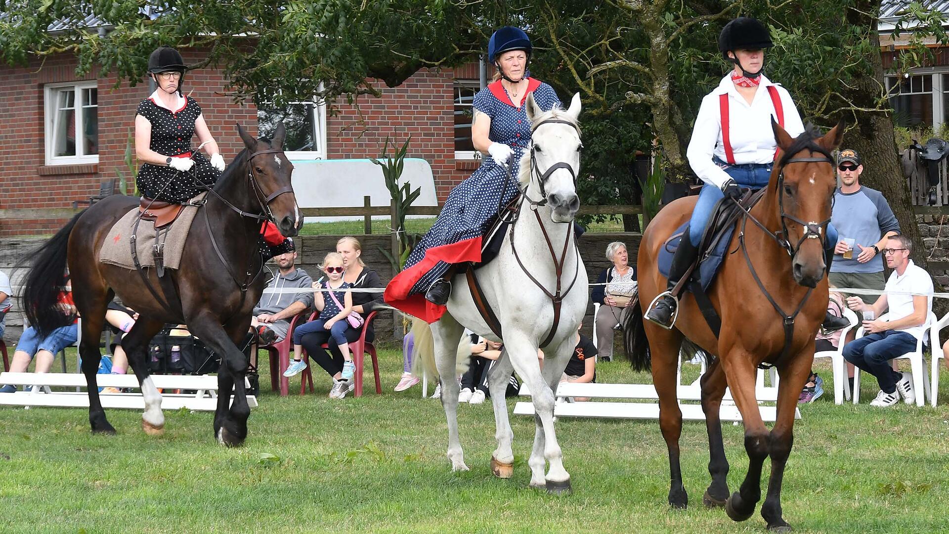 Drei Frauen in verschiedenen nostalgischen Outfits reiten auf ihren Pferden bei einer Vorführung auf dem Midlumer Markt. Im Hintergrund verfolgen Zuschauer das Geschehen vom Rand der Reitbahn.