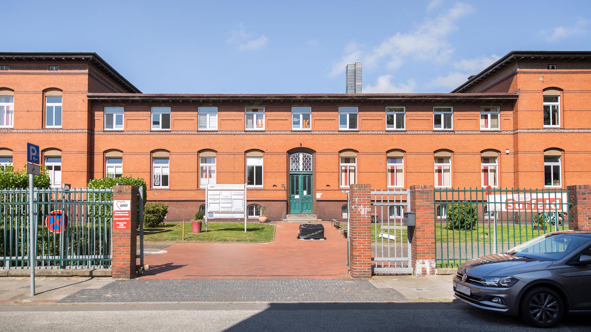 rotes Backsteingebäude mit grüner Tür in einer parkähnlichen Anlage hinter einem Zaun
