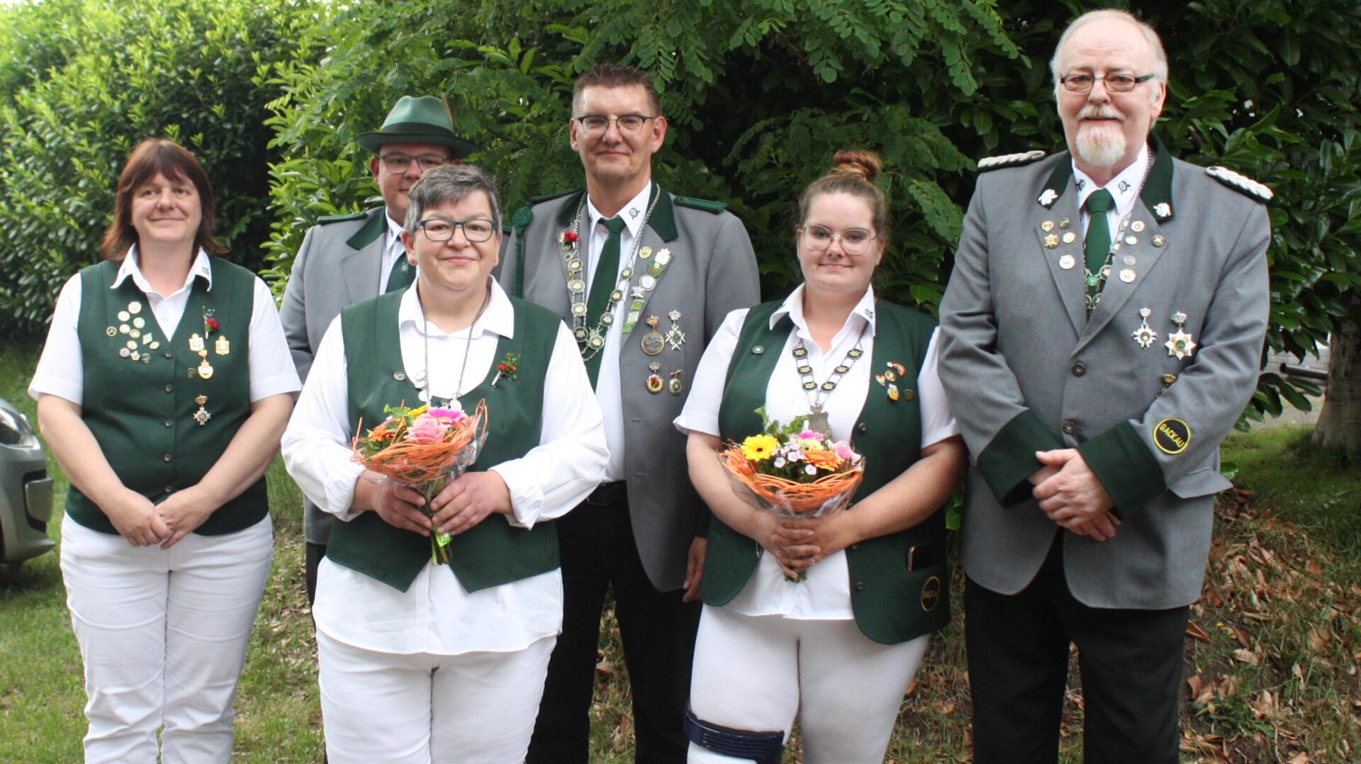 **ALT-Text:**
Sechs Mitglieder des Schützenvereins Gackau stehen in Uniform nebeneinander und lächeln in die Kamera. Zwei Frauen in der vorderen Reihe halten Blumensträuße. Alle tragen Schützenabzeichen und Ordensketten.