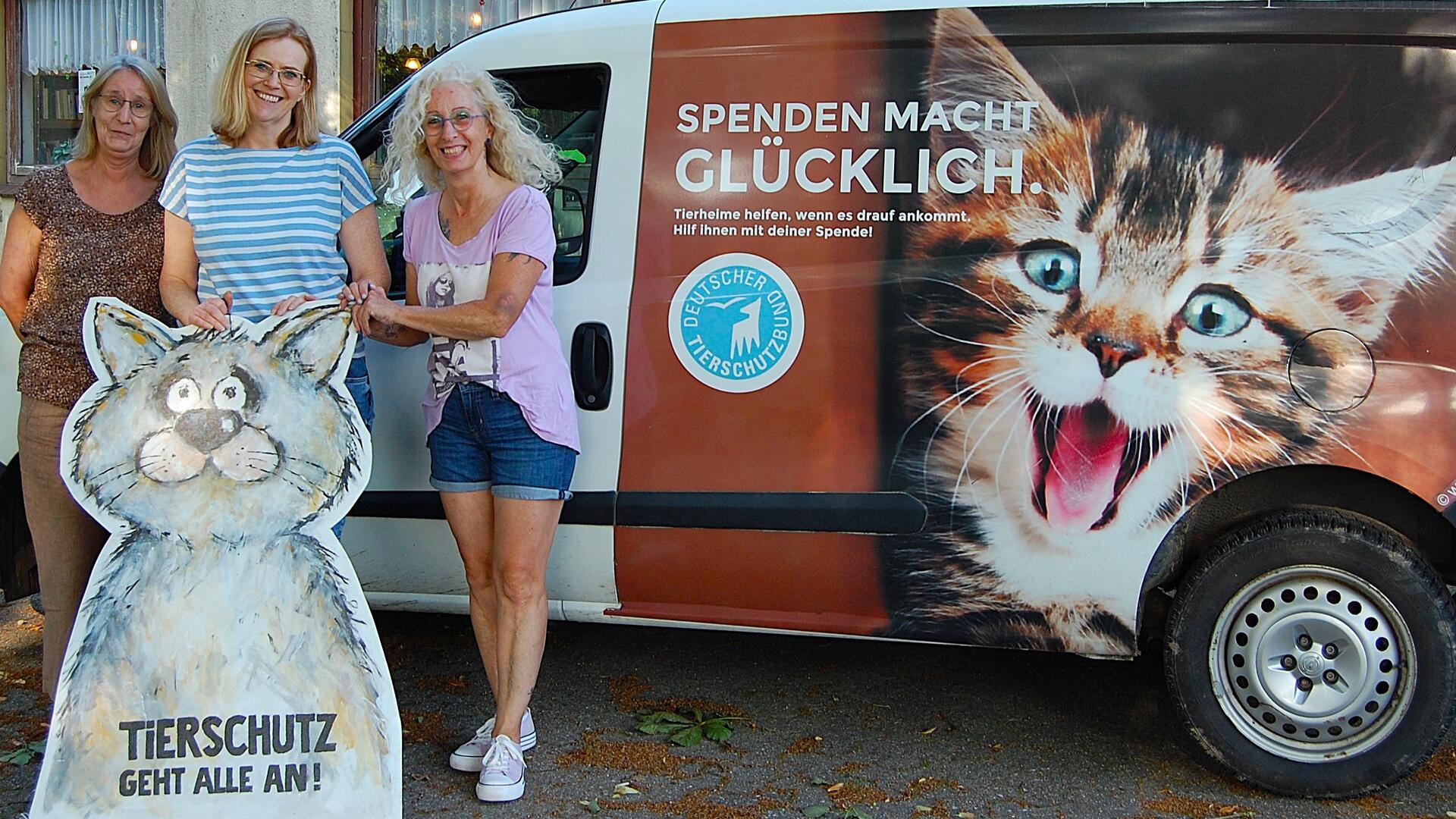 Drei Frauen stehen vor einem Transporter mit der Aufschrift „Spenden macht glücklich“ und einem großen Katzenmotiv. Sie halten ein gemaltes Pappschild mit der Aufschrift „Tierschutz geht alle an!“ in Form einer Katze.