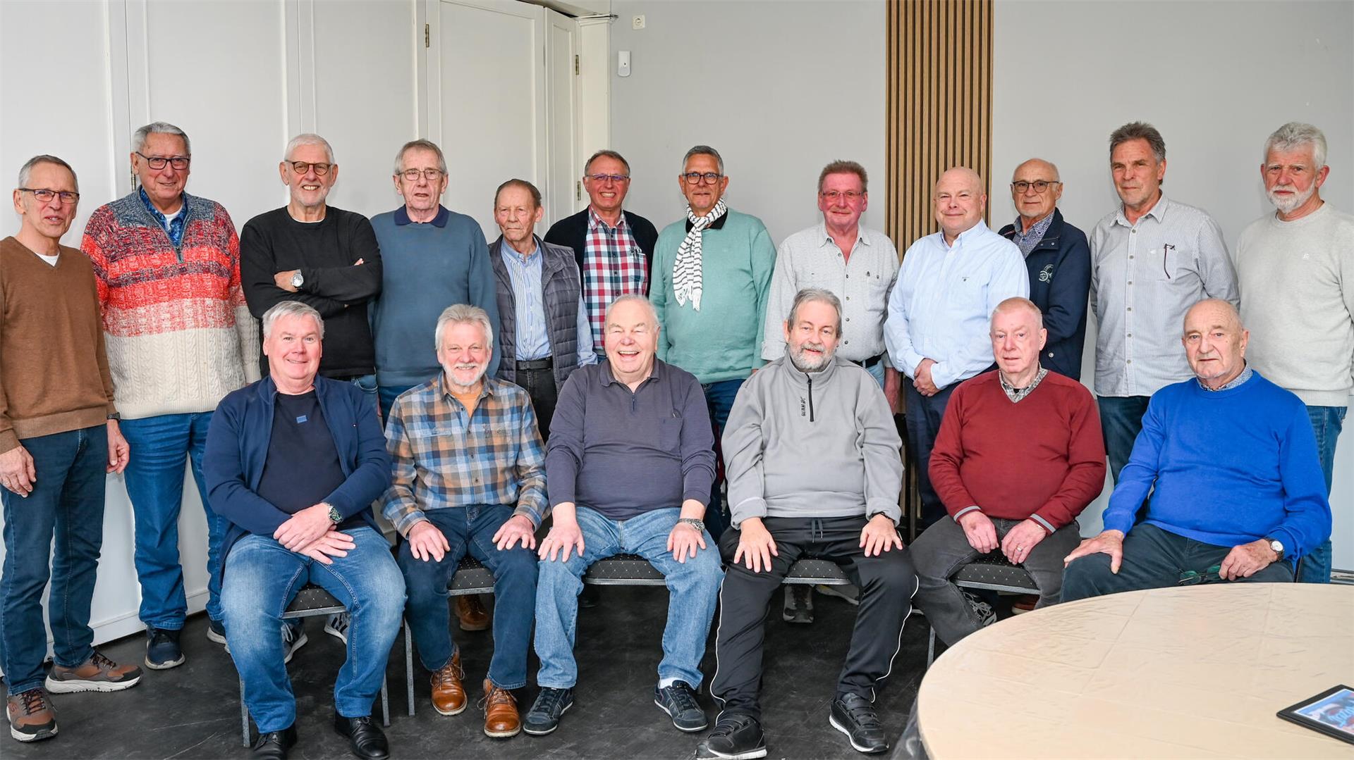 Gruppenfoto von 18 älteren Männern in Freizeitkleidung, aufgenommen in einem hellen Raum. Einige sitzen, andere stehen dahinter. 