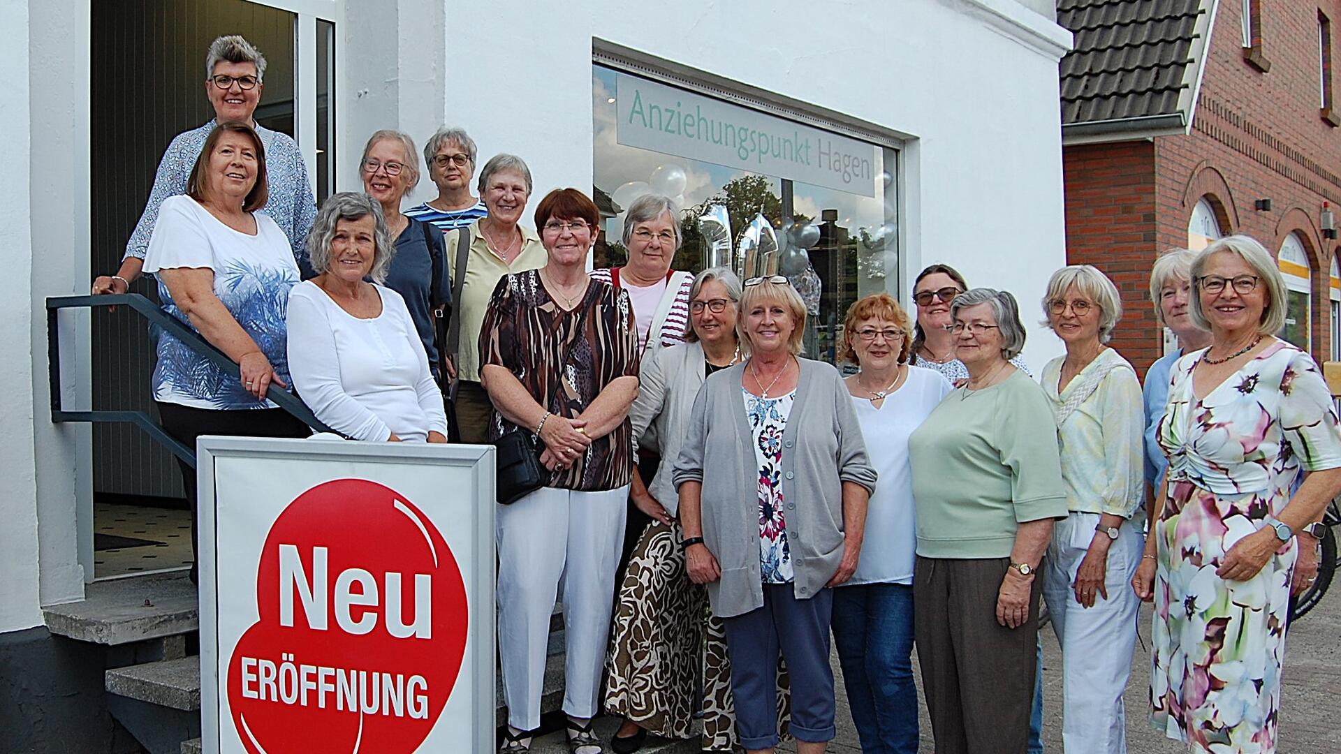 Frauen aus dem Team der Kleiderkammer „Anziehungspunkt“ in Hagen stehen lächelnd vor dem Eingang des Ladens neben einem Schild mit der Aufschrift „Neu-Eröffnung“.