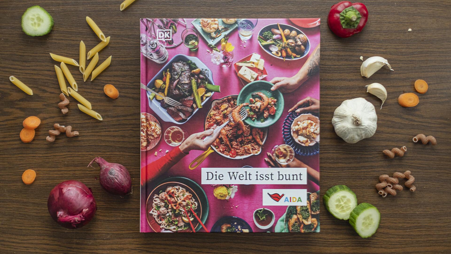 Das Kochbuch "Die Welt isst bunt" auf einem Holztisch, umgeben von Gemüse und Kochzutaten. 