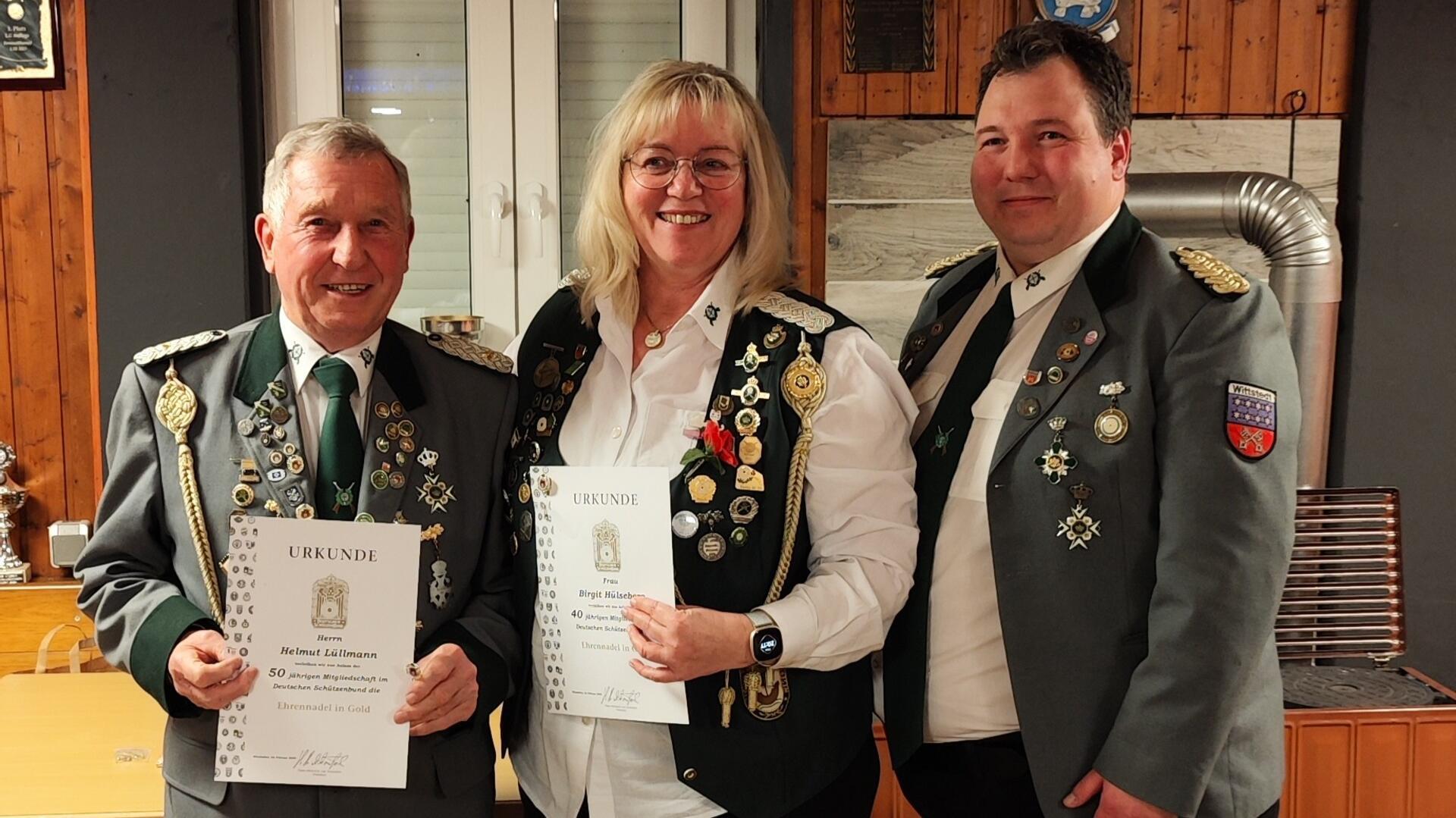 Drei Personen in traditionellen Schützenuniformen stehen in einem Raum mit Holzwänden. Sie tragen zahlreiche Abzeichen und Orden an ihren Jacken und Westen. Die beiden Personen links, ein älterer Mann und eine Frau mit blonden Haaren, halten jeweils eine Urkunde.