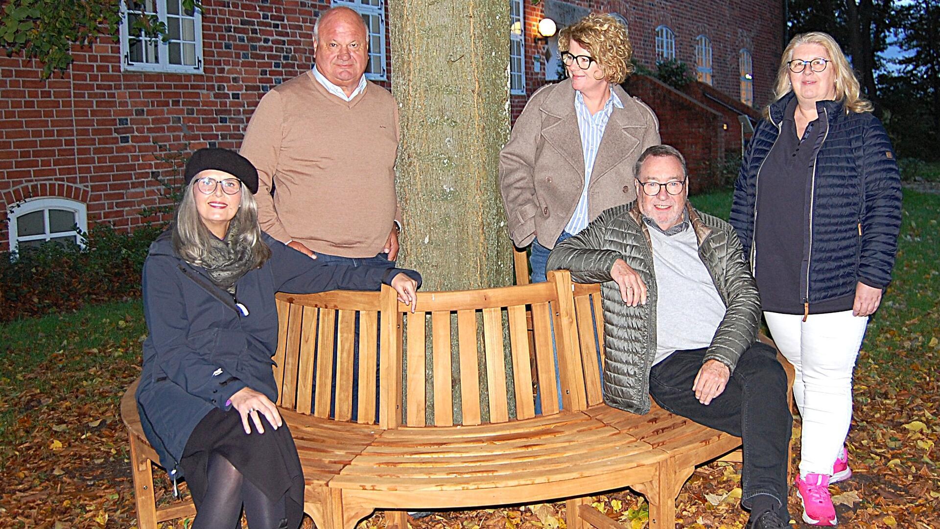 Fünf Personen stehen und sitzen um eine halbrunde Holz-Baumbank im herbstlich gefärbten Burghof. Im Hintergrund ist ein historisches Backsteingebäude zu sehen.