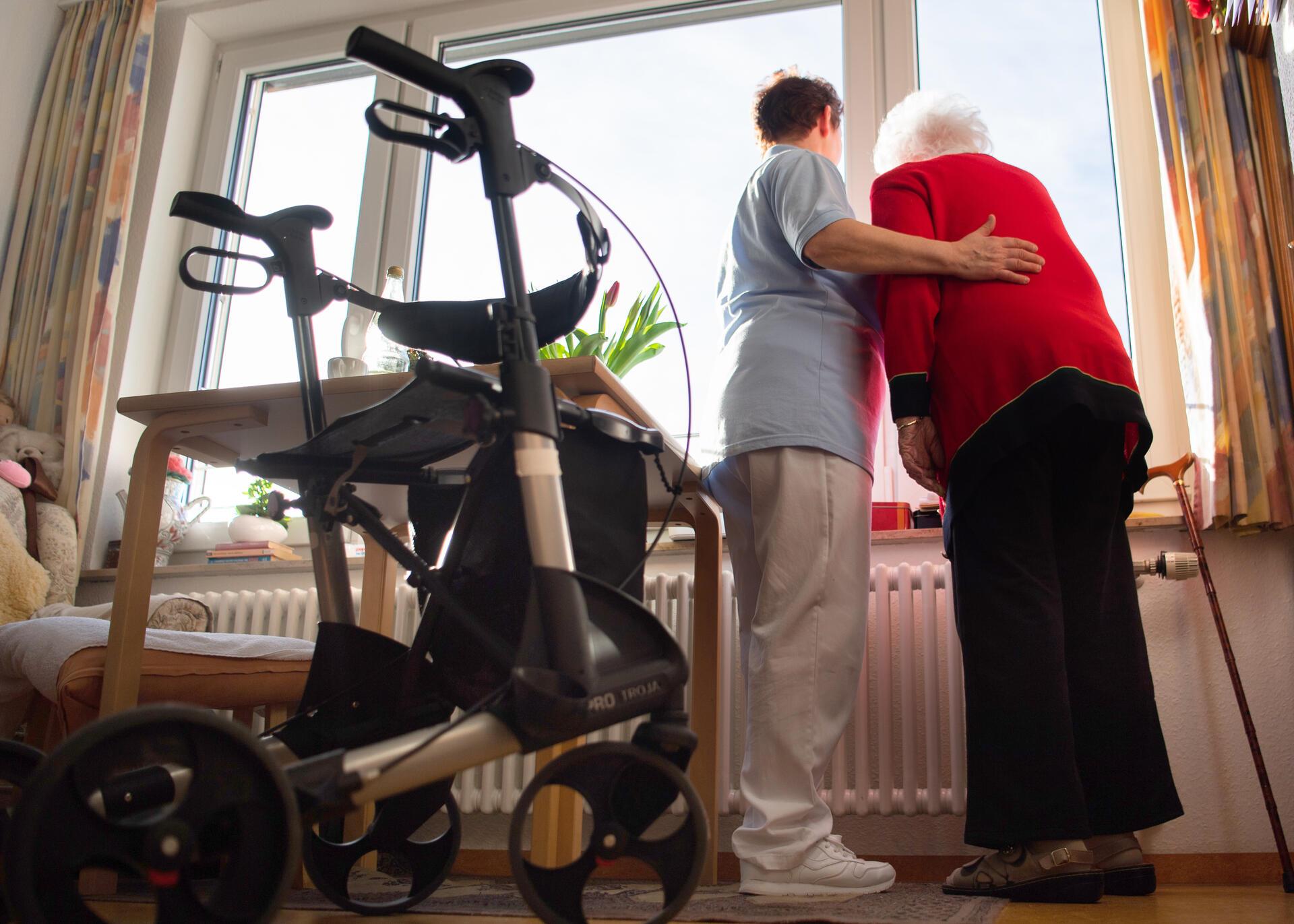 Eine Pflegerin (links) und eine Heimbewohnerin schauen zusammen aus einem Fenster im Wohnbereich eines Pflegeheims. 