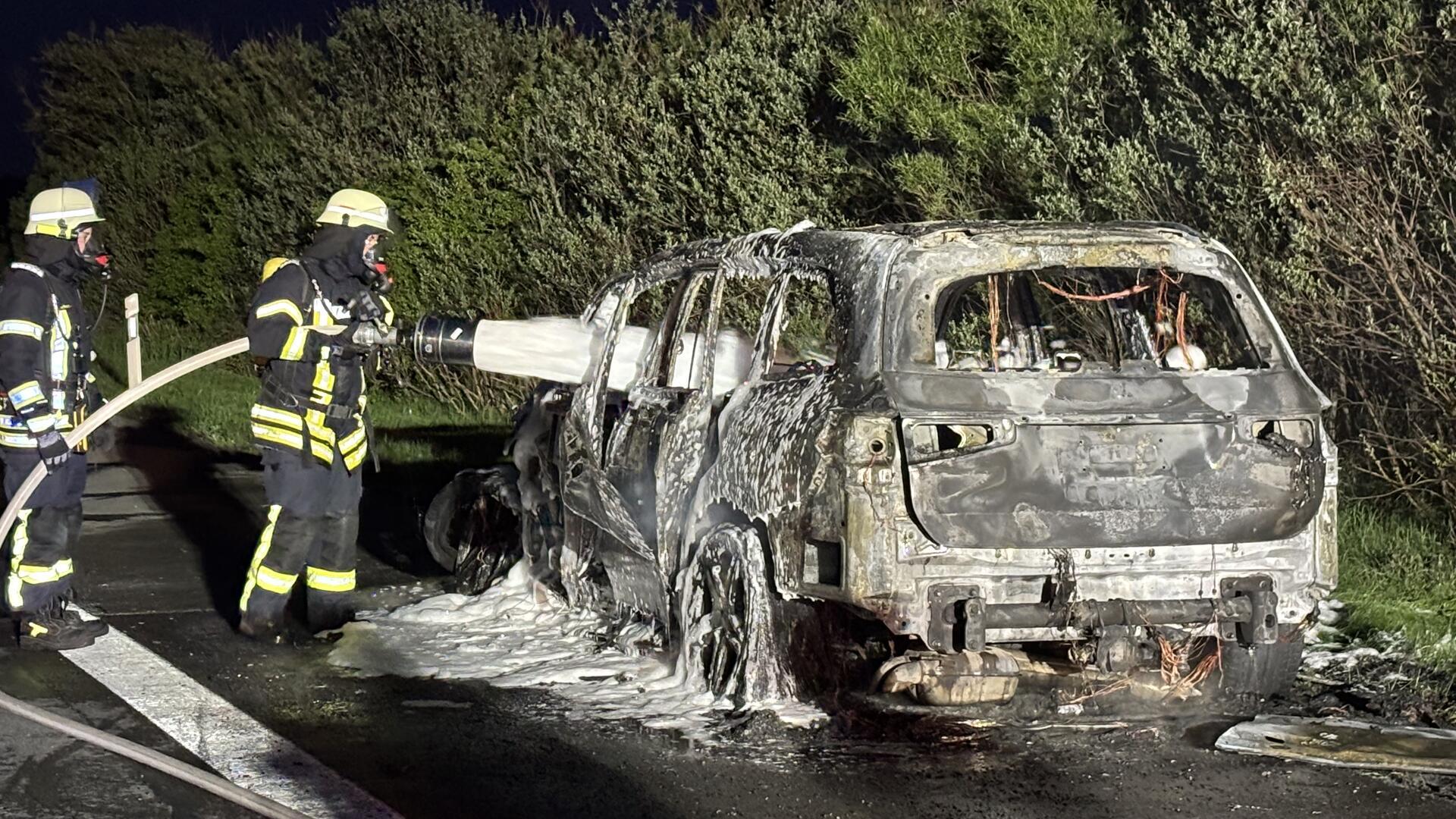 Zwei Feuerwehrleute stehen neben einem ausgebrannten Auto und besprühen es mit Löschschaum.