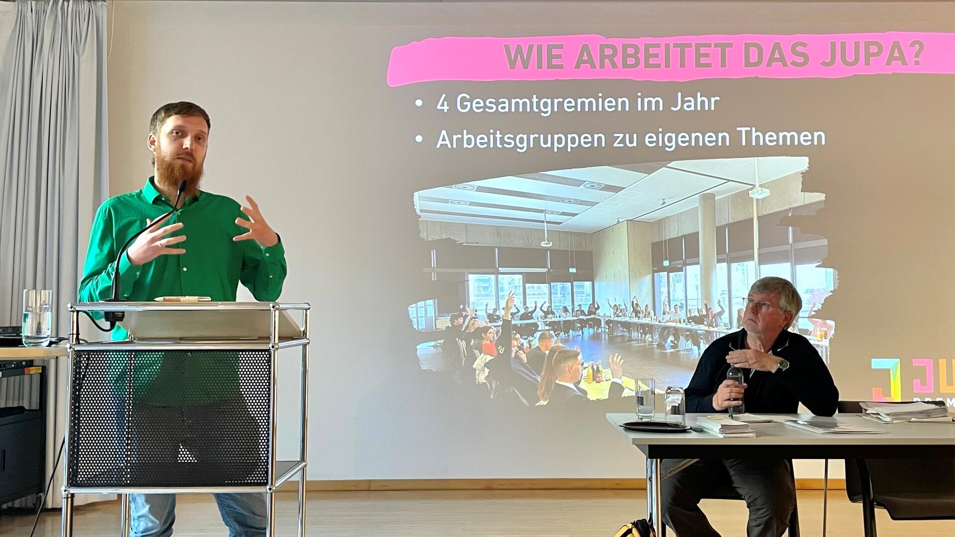 Ein Mann mit grünem Hemd steht an einem Rednerpult. Er spricht. Rechts neben ihm sitzt ein Mann mit schwarzem Oberteil auf einem Stuhl und an einem Tisch. Hinter den Männern macht eine Präsentation auf das Jugendparlament aufmerksam. Es geht um die Frage, wie das Gremium arbeitet.