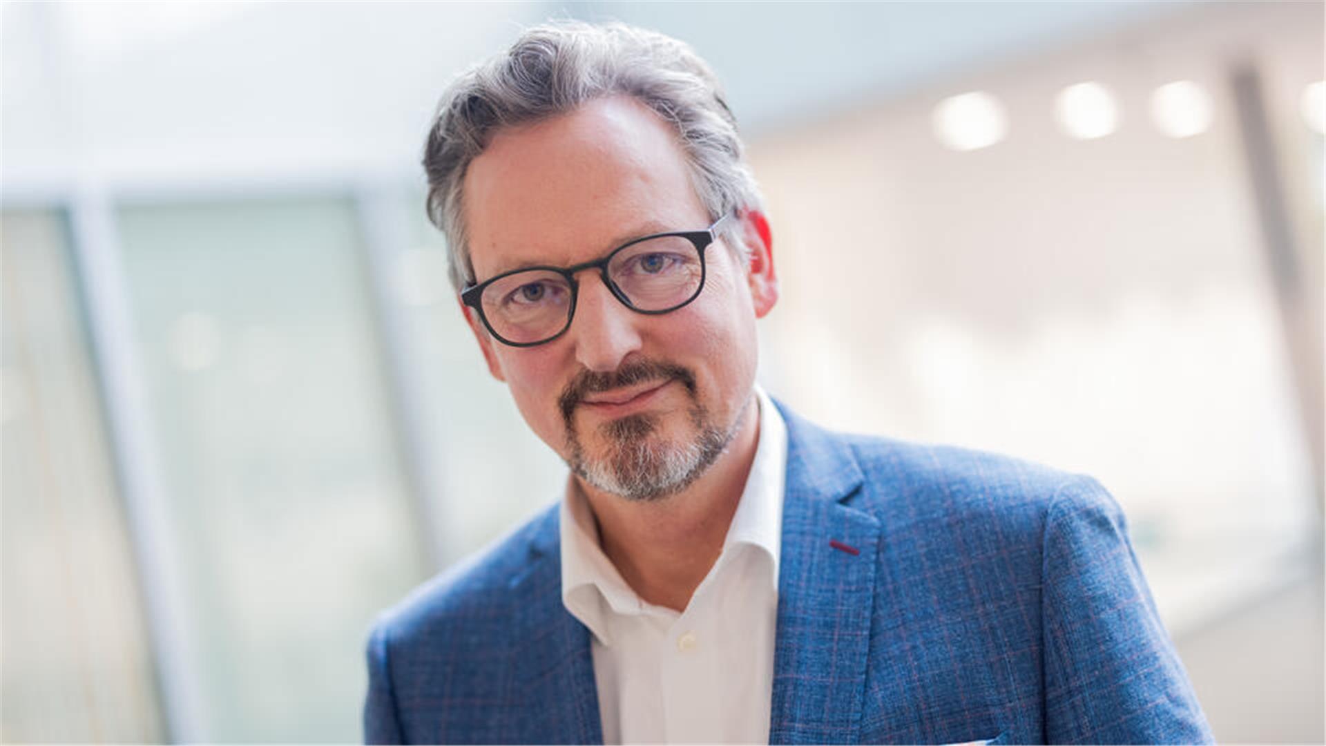 Mann mit grauen Haaren, Bart und Brille lächelt in die Kamera. Ein heller verschwommener Hintergrund.
